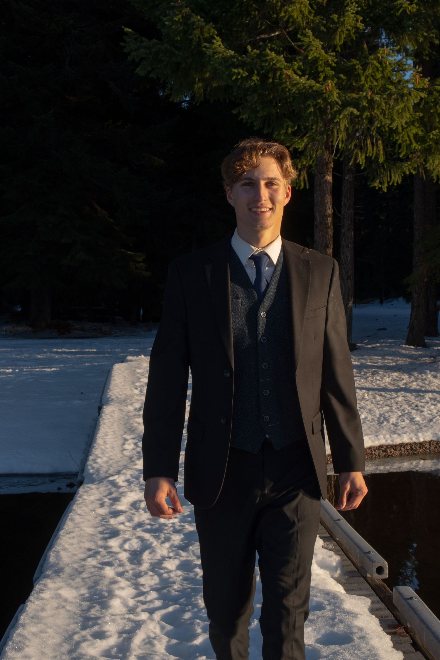 Young groom smiling in black suit and tie standing on snowy dock with evergreen trees behind.