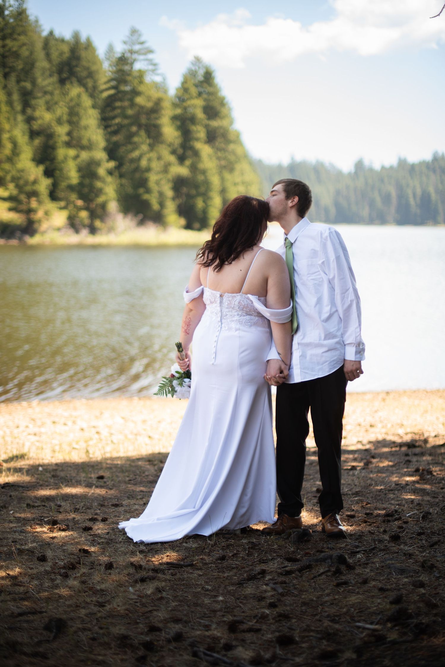 Bride and groom share a kiss by a serene lake surrounded by pine trees on their wedding day.