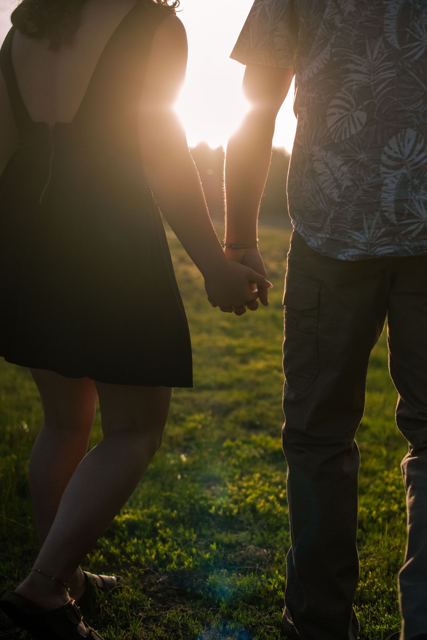 A couple holds hands in silhouette against a stunning golden sunset on a green grassy field.