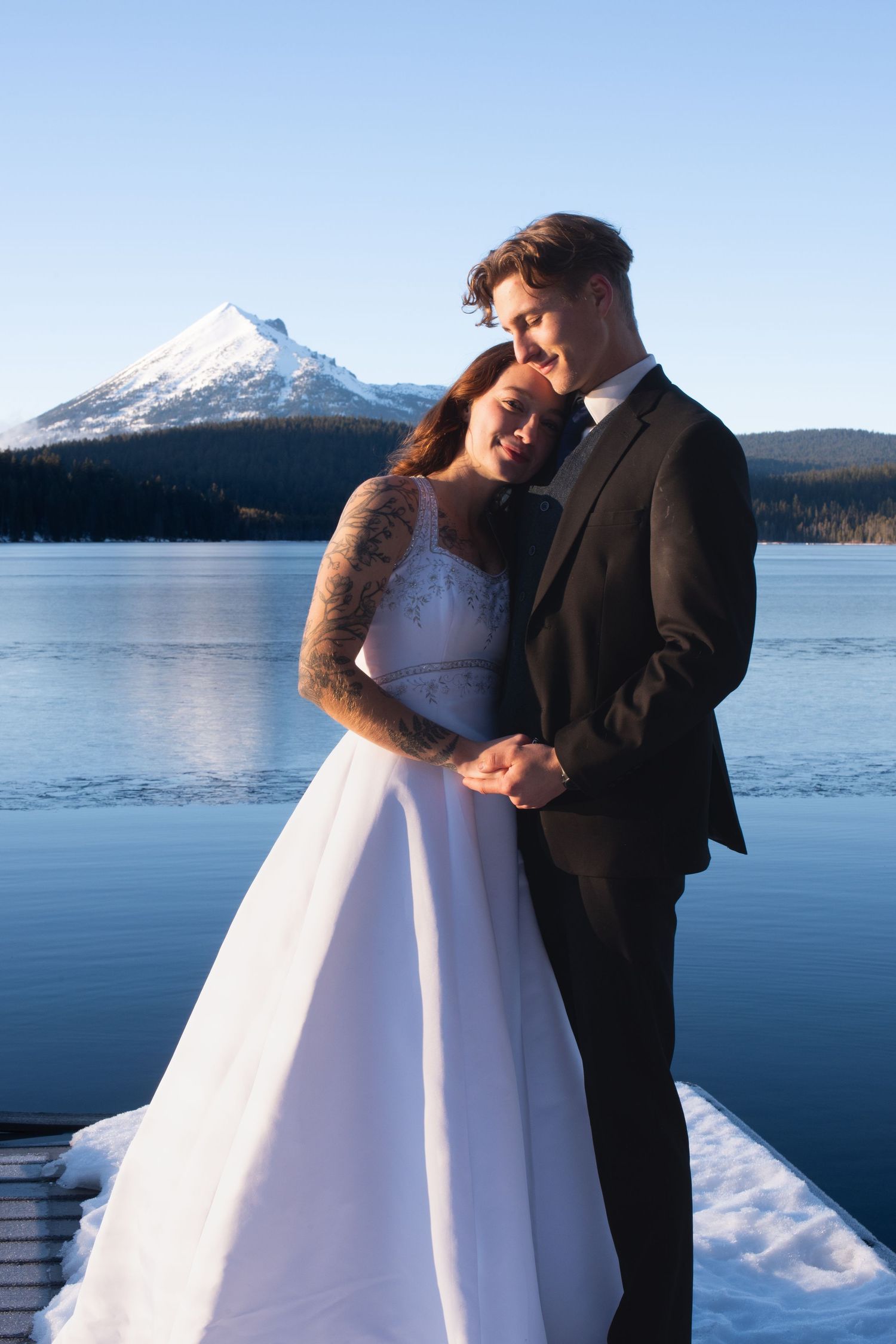 Elegant bride and groom embrace on a snowy dock with a stunning snow-capped mountain and lake backdrop.