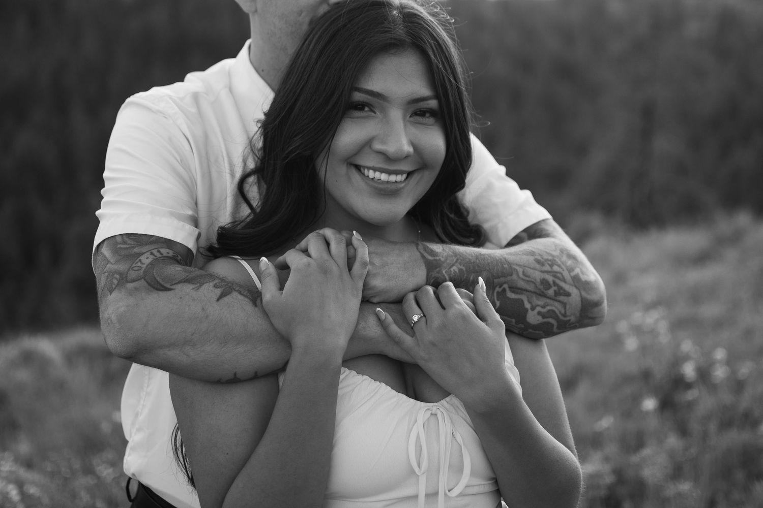 Black and white photo of a smiling woman being embraced from behind by a tattooed man outdoors.