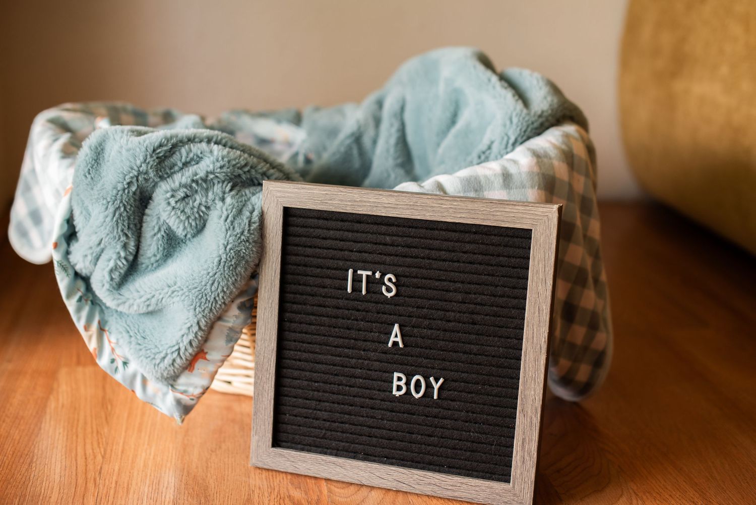 Letter board sign reading It's A Boy displayed beside a teal baby blanket in a wicker basket.