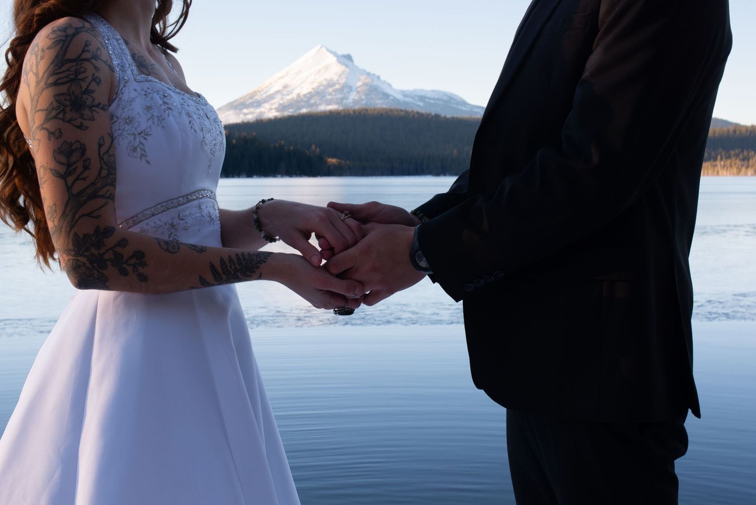 Close-up of tattooed bride and groom exchanging rings with mountain lake in background.