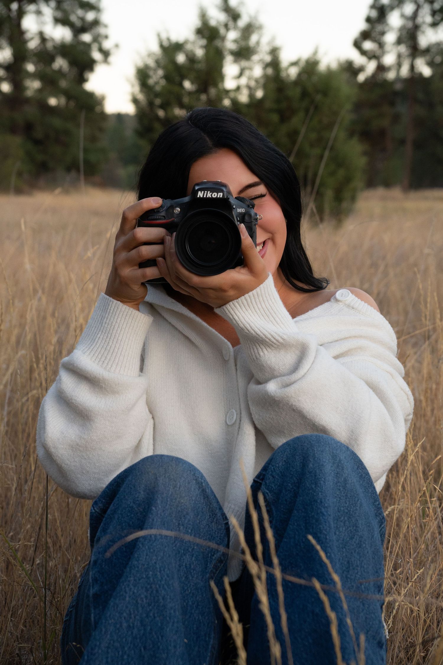 Woman in white sweater and jeans sitting in a field, holding a Nikon DSLR camera up to her face.
