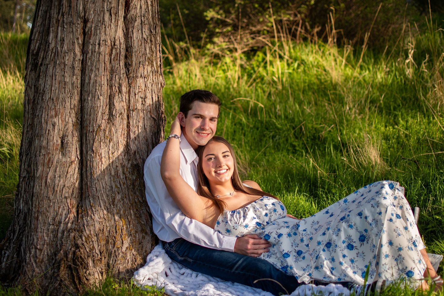 Couple sitting together against a tree trunk in a sunny green meadow, woman in floral dress smiling warmly.