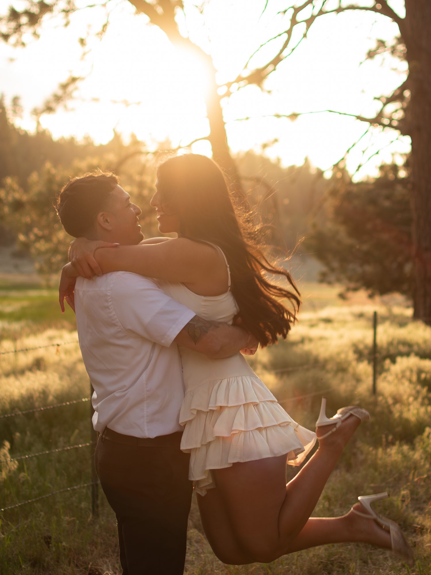 Couple embracing at golden hour in a sunlit meadow, man lifting woman in white ruffled dress near pine trees.