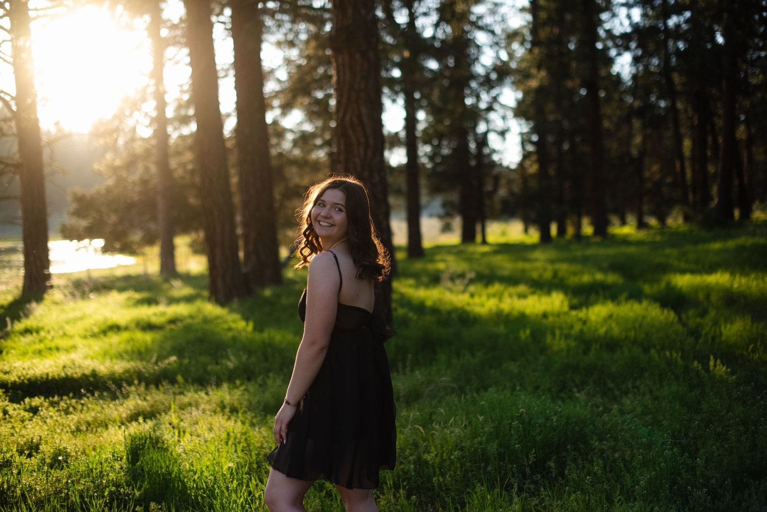 Young woman in black dress poses in a sunlit forest meadow surrounded by tall pine trees.