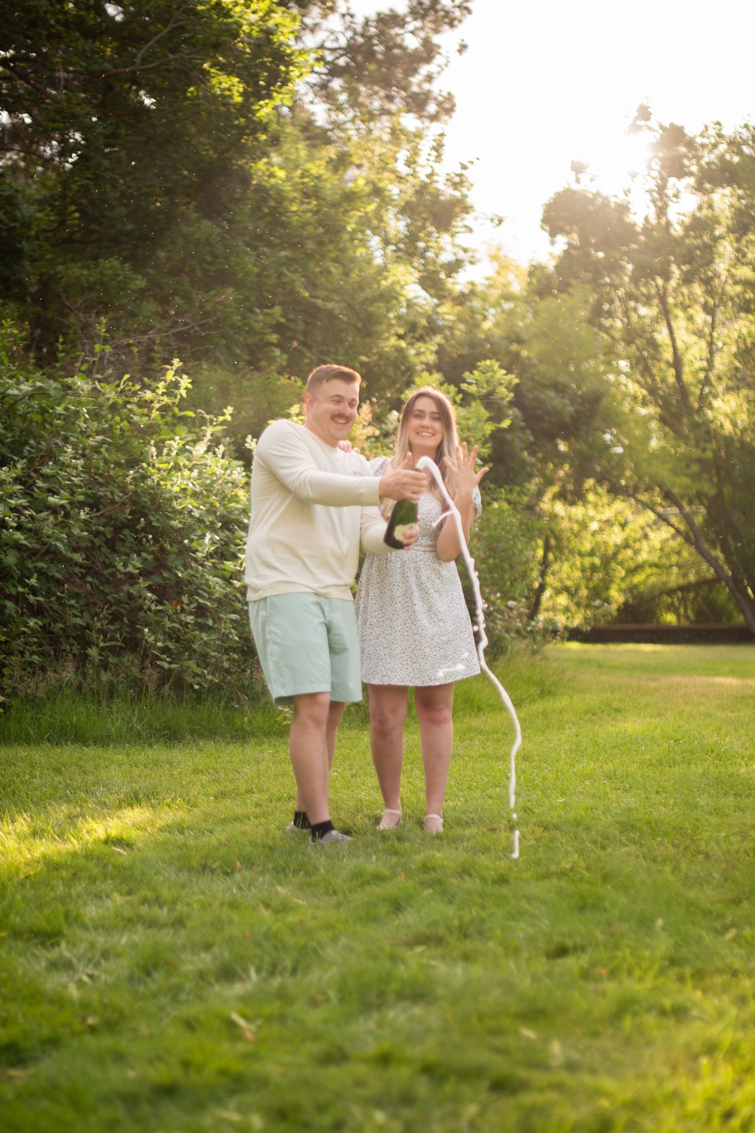 Excited couple popping champagne on a green lawn at sunset, woman displaying engagement ring with joy.