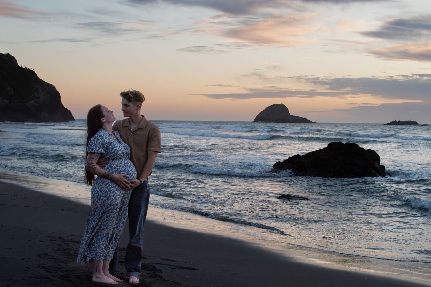 Pregnant couple sharing a tender moment on a black sand beach at sunset.