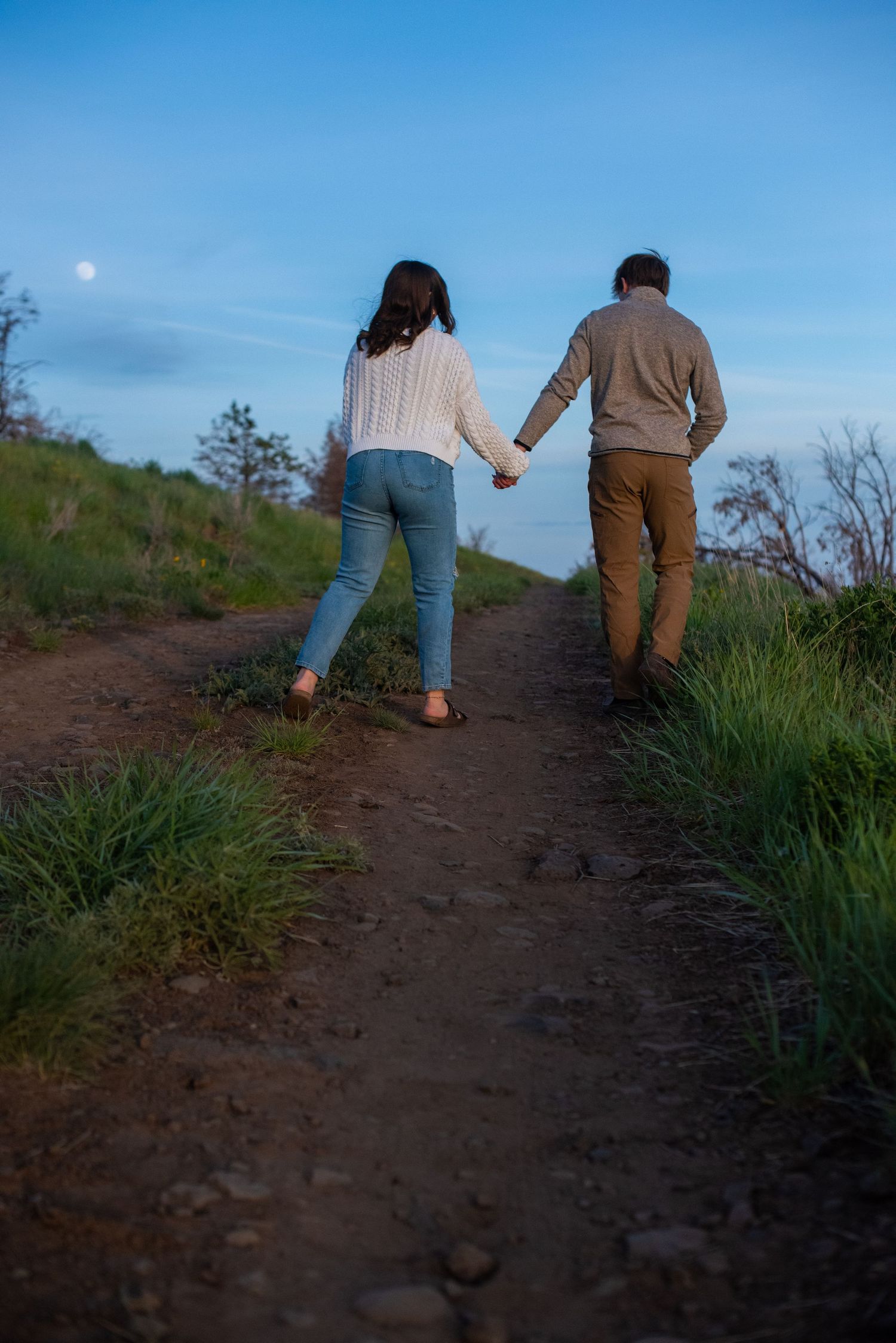 Couple holding hands walking along a dirt path at dusk under a blue sky with visible moon.