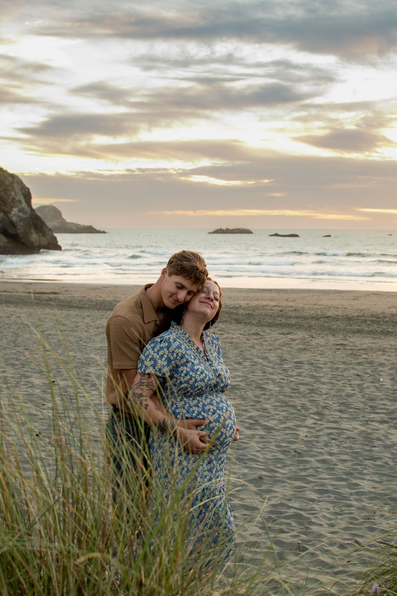 Couple sharing a tender embrace on a sandy beach at sunset, with the pregnant woman cradling her bump.