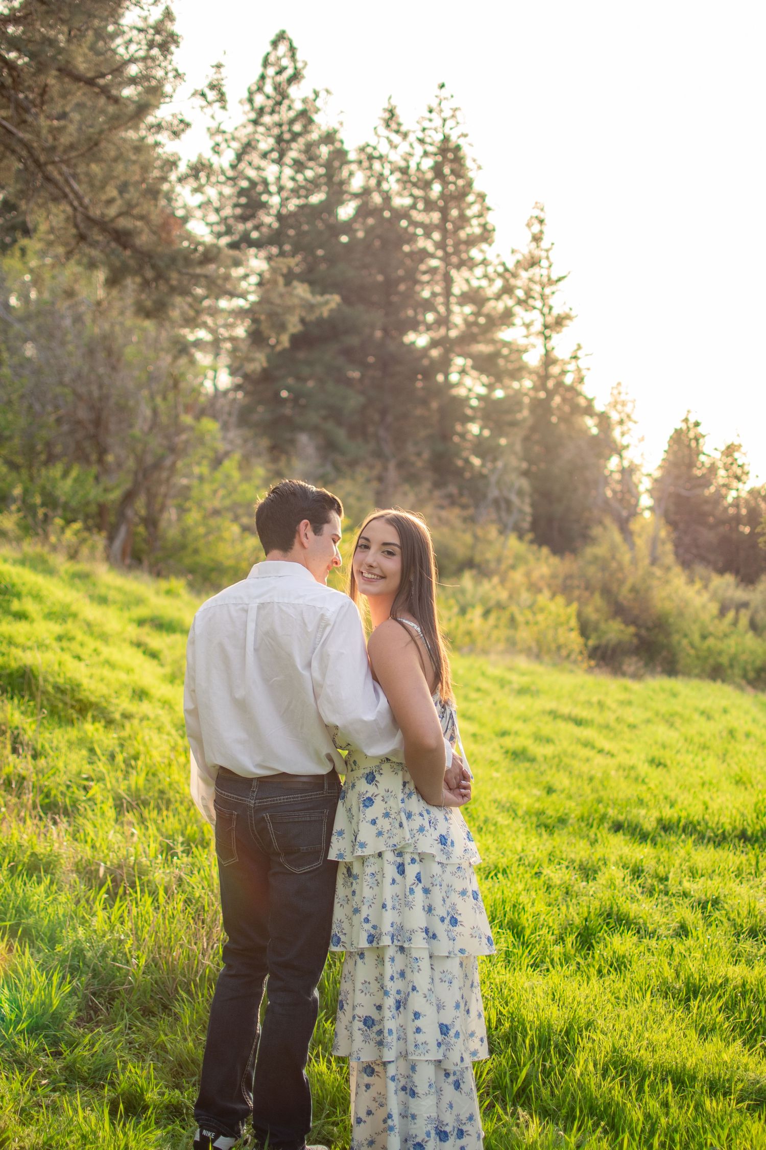 Couple embracing in sunlit green meadow with tall pine trees, woman in floral dress smiling over shoulder.
