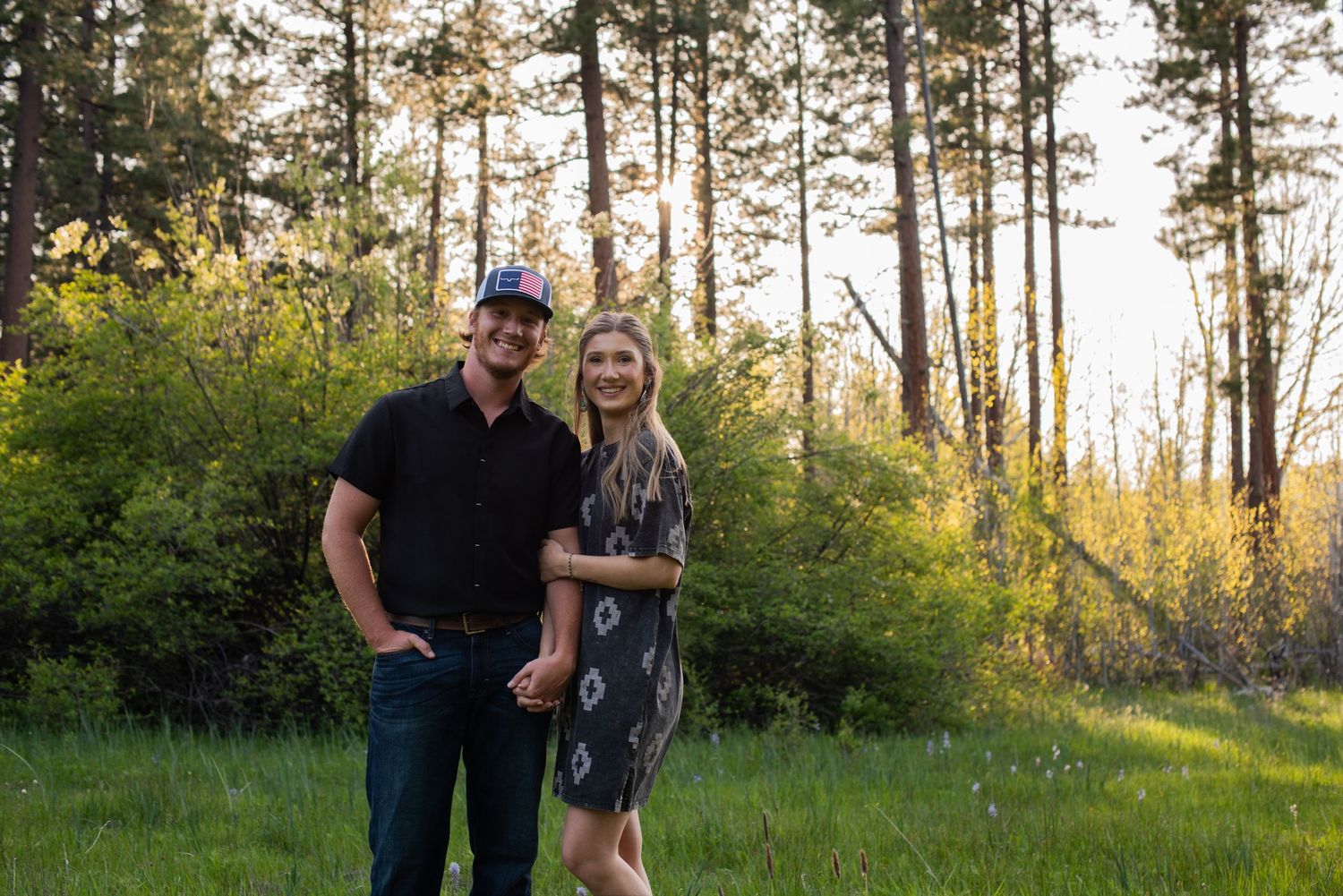 A smiling couple holds hands in a lush green forest clearing at golden hour, man in black shirt and cap, woman in floral dress.