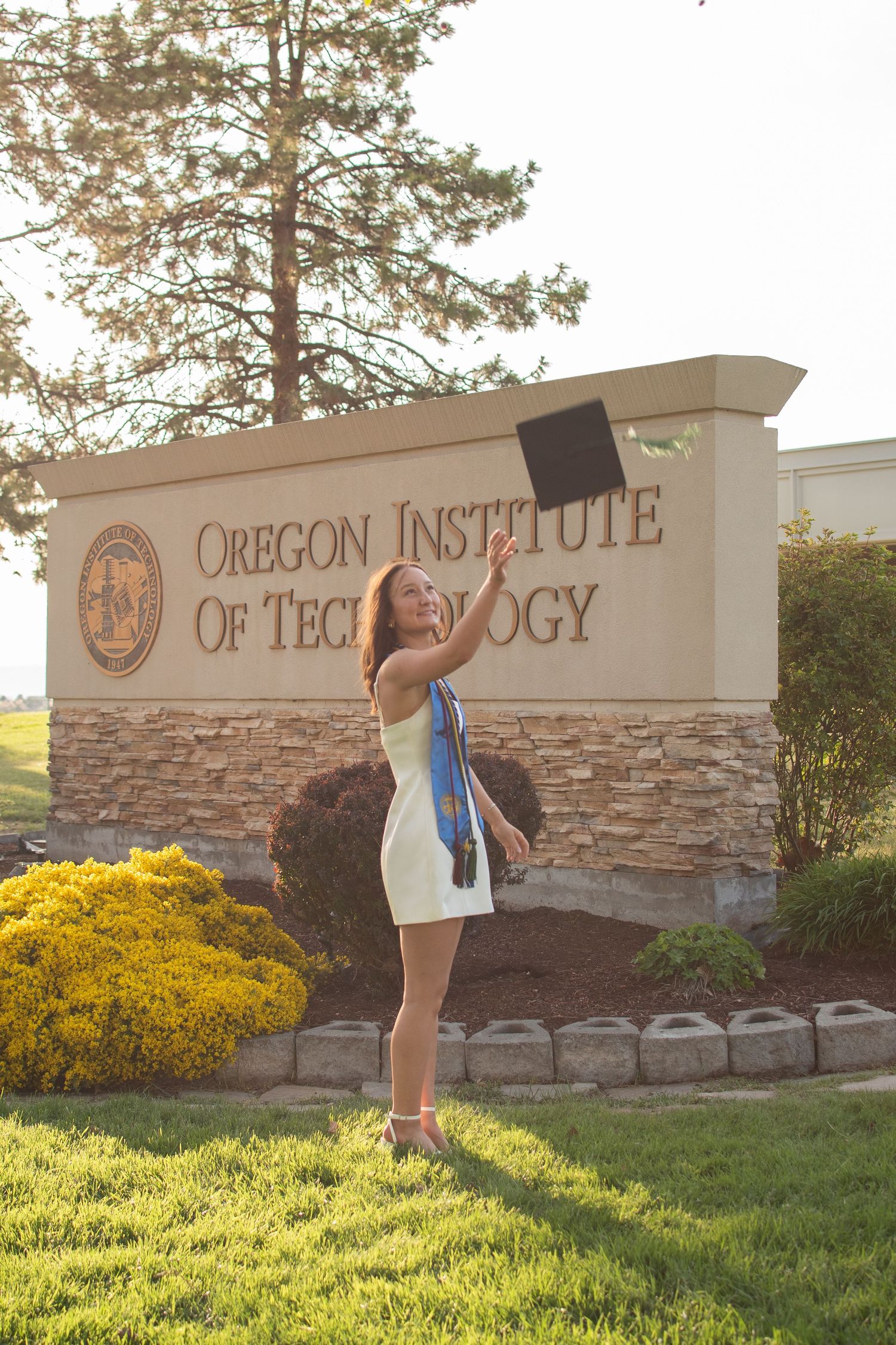 Graduate celebrates in front of Oregon Institute of Technology sign, tossing cap in white dress.
