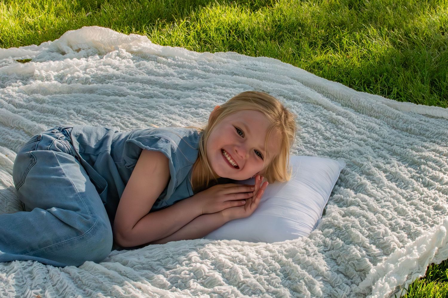 Smiling young girl lying on a white fluffy blanket with a pillow on green grass outdoors.