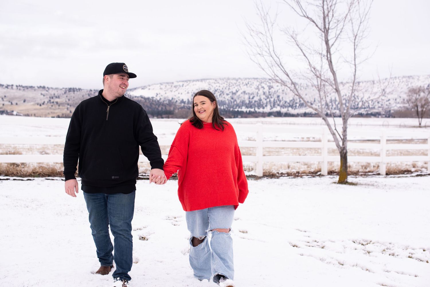 Couple holding hands walking in snowy winter landscape, man in black jacket and woman in red sweater.