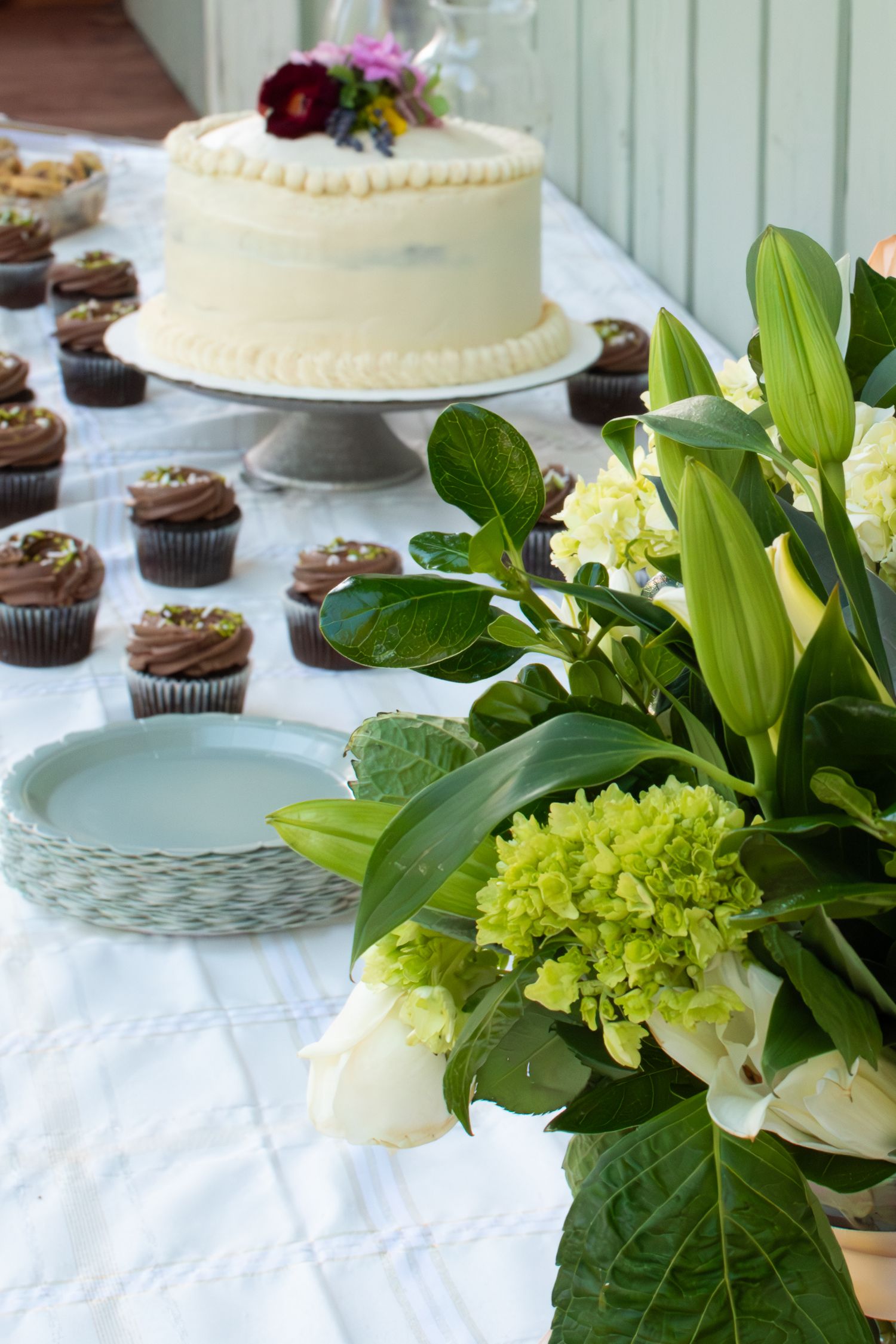 Elegant dessert table with white frosted cake, chocolate cupcakes, green hydrangea floral arrangement, and blue decorative plate.