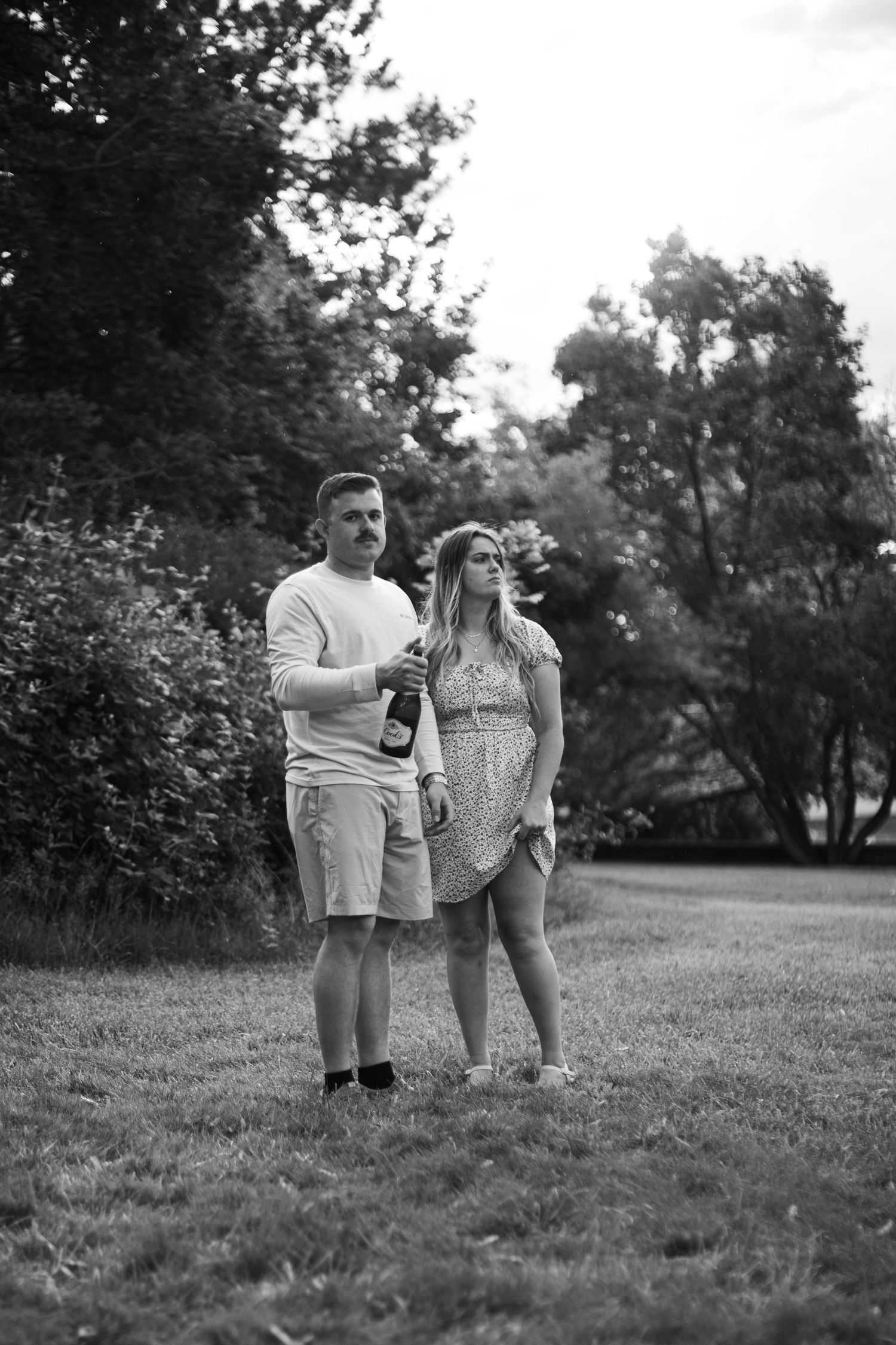 Black and white photo of a couple standing together on a grassy lawn holding a bottle of champagne.