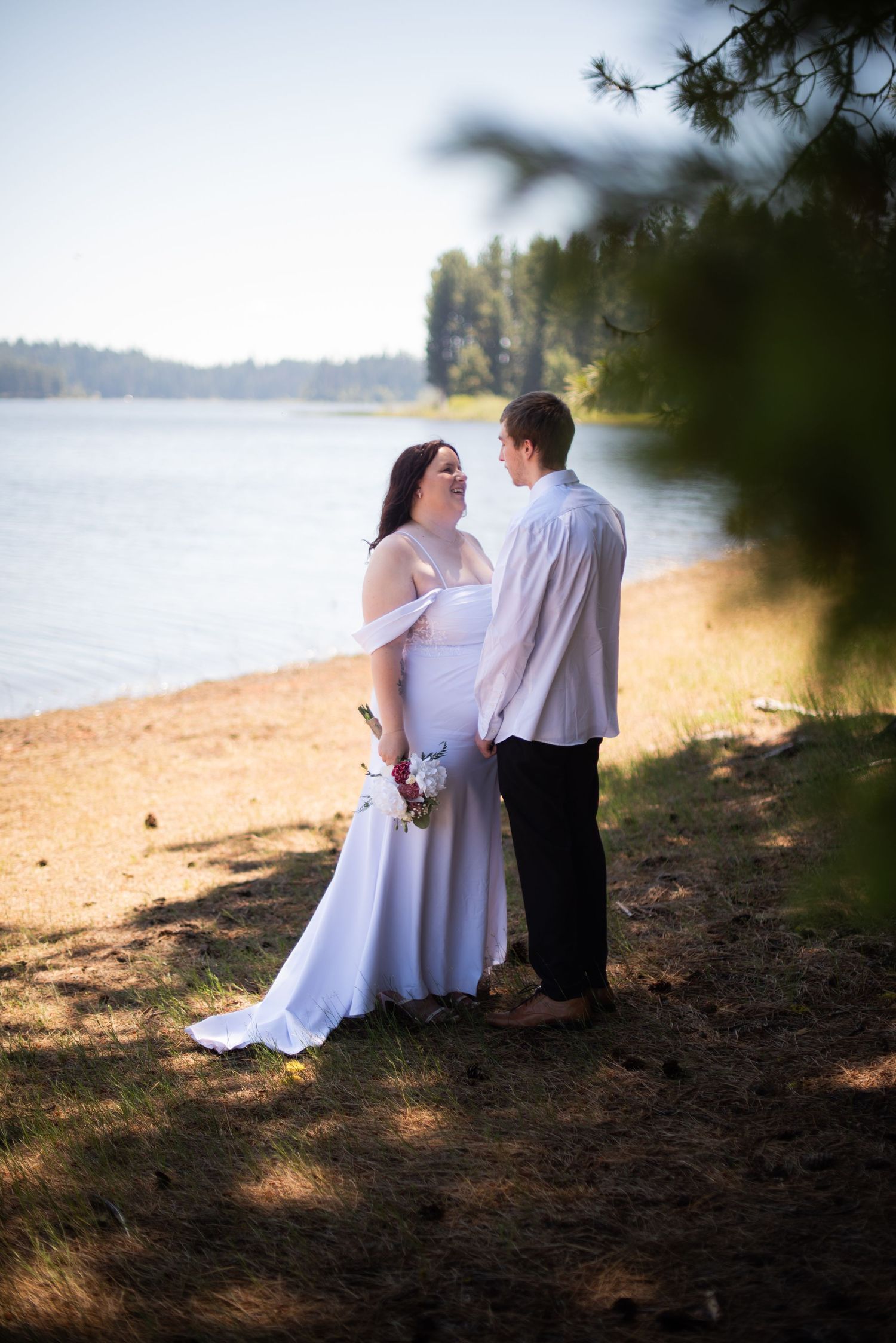 Bride in white gown and groom share a moment on a sunny lakeside beach surrounded by pine trees.
