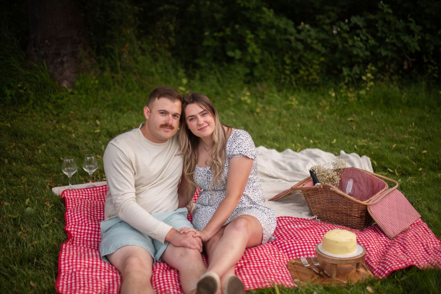 Couple with fake mustaches sitting closely together on a red checkered picnic blanket in a green park.