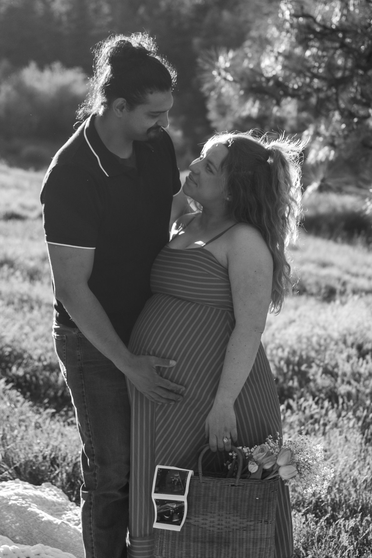 Black and white photo of a pregnant couple embracing outdoors, woman holding ultrasound photos and a basket.