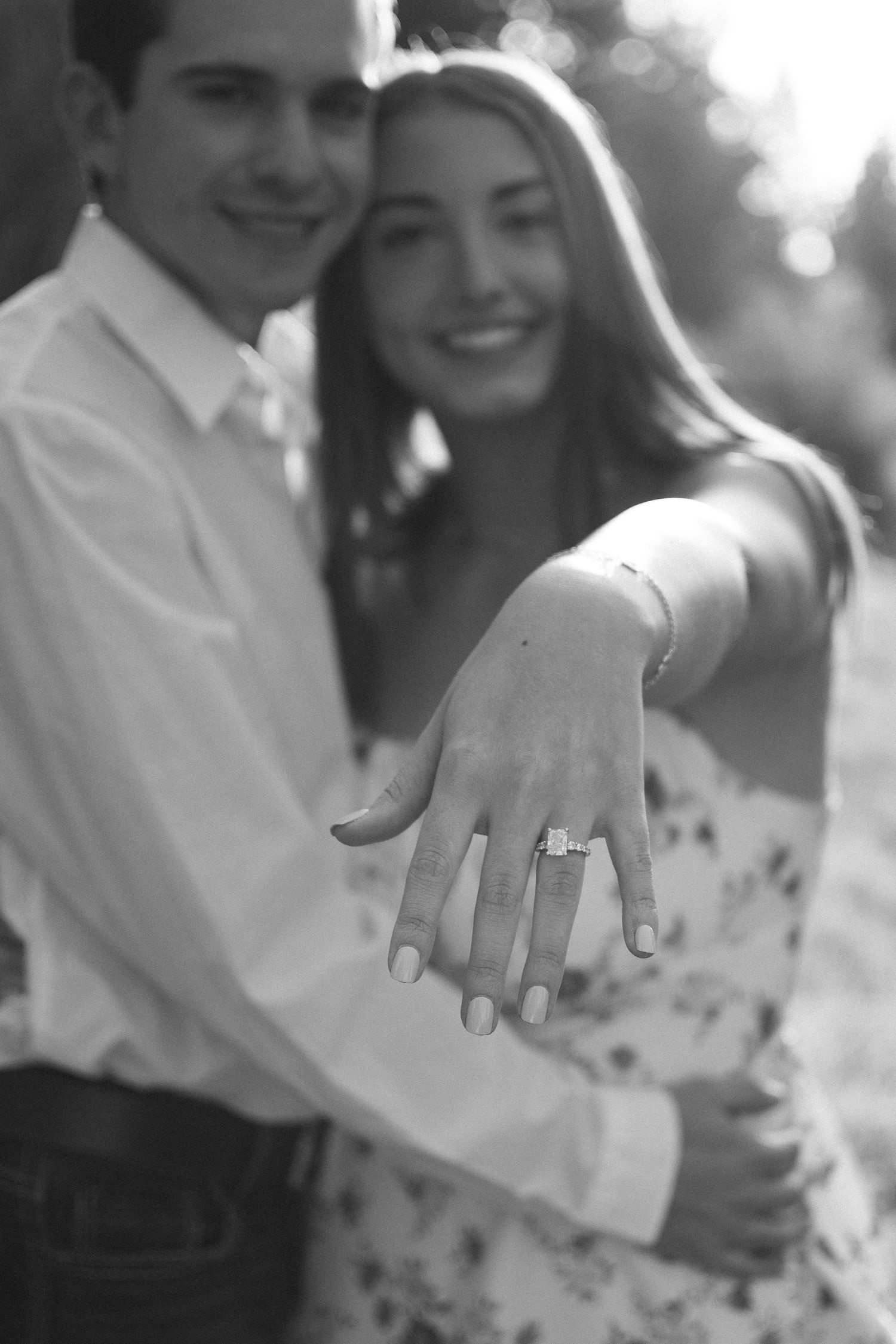 Black and white photo of smiling couple showing off engagement ring outdoors.