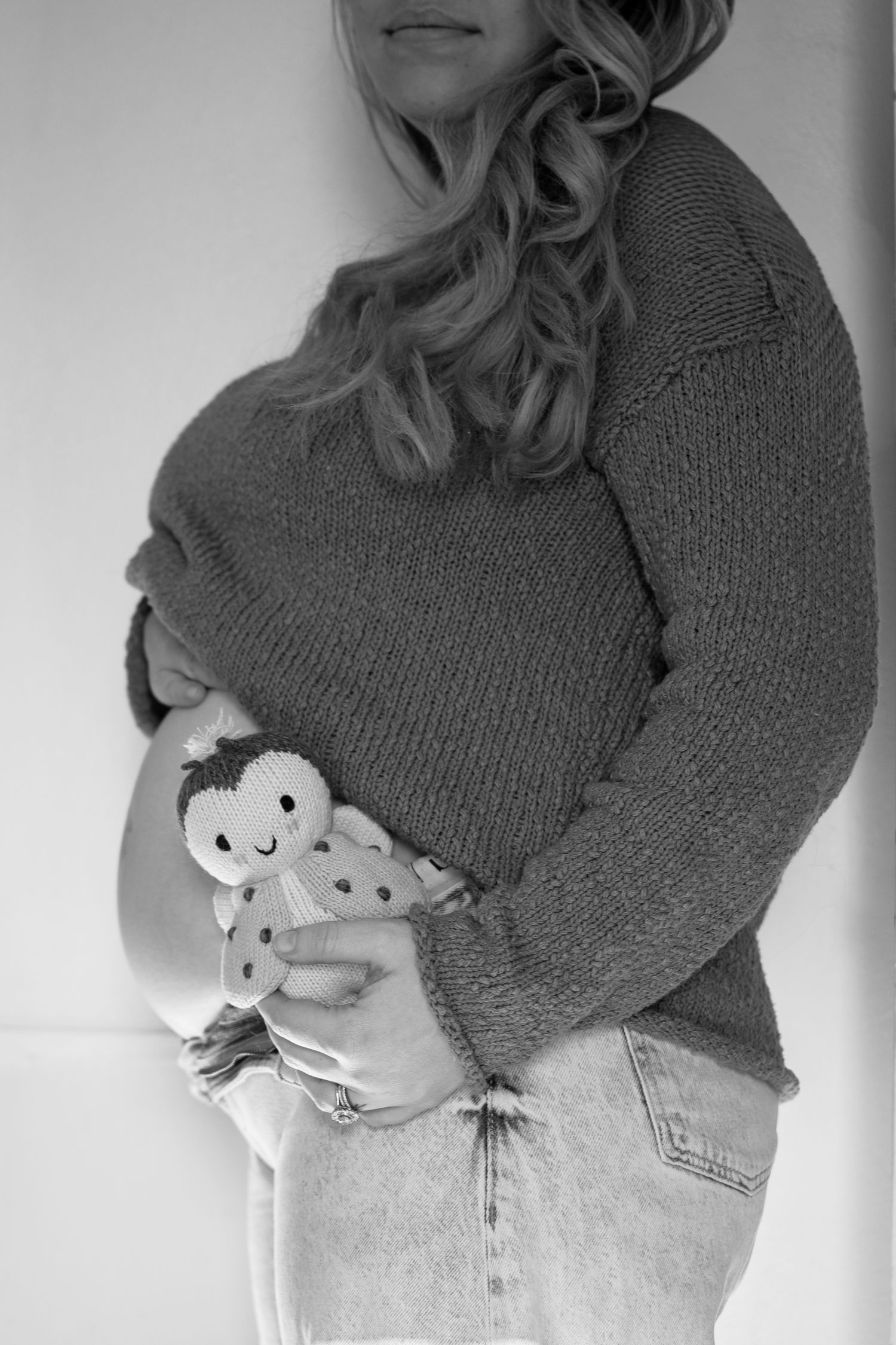 Black and white photo of a pregnant woman in a knit sweater holding a small knitted doll against her baby bump.