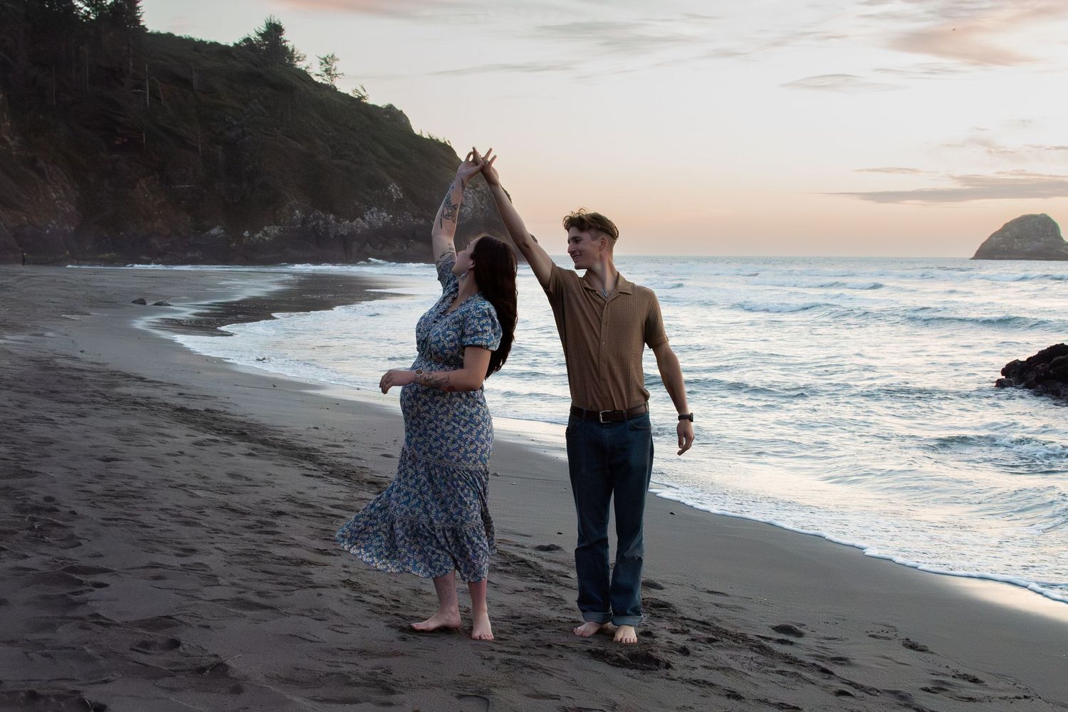 Couple dancing on a beach at sunset, woman in floral dress, man in brown shirt, ocean waves and green cliffs behind.
