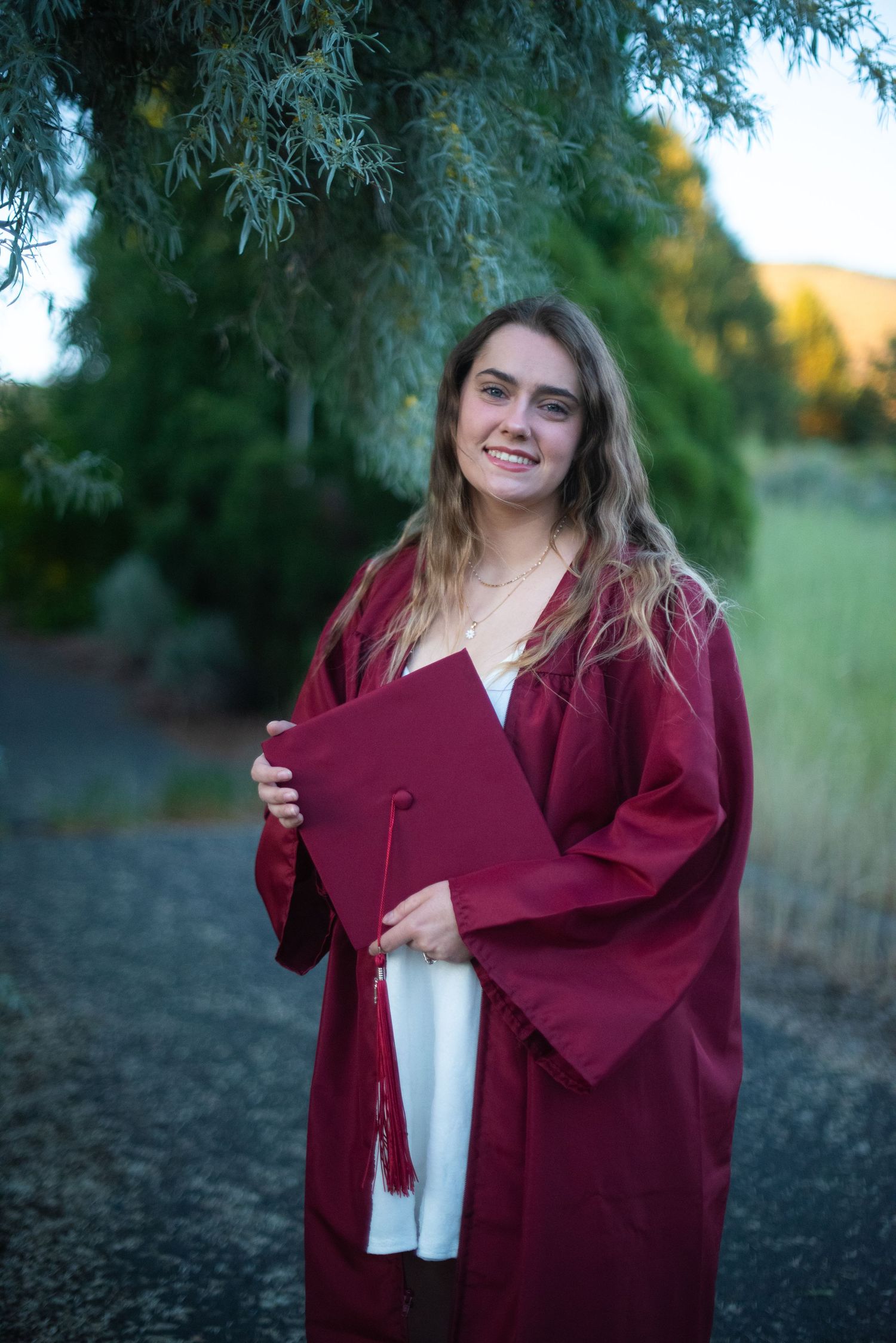 Smiling young woman in red graduation cap and gown holding her mortarboard outdoors.