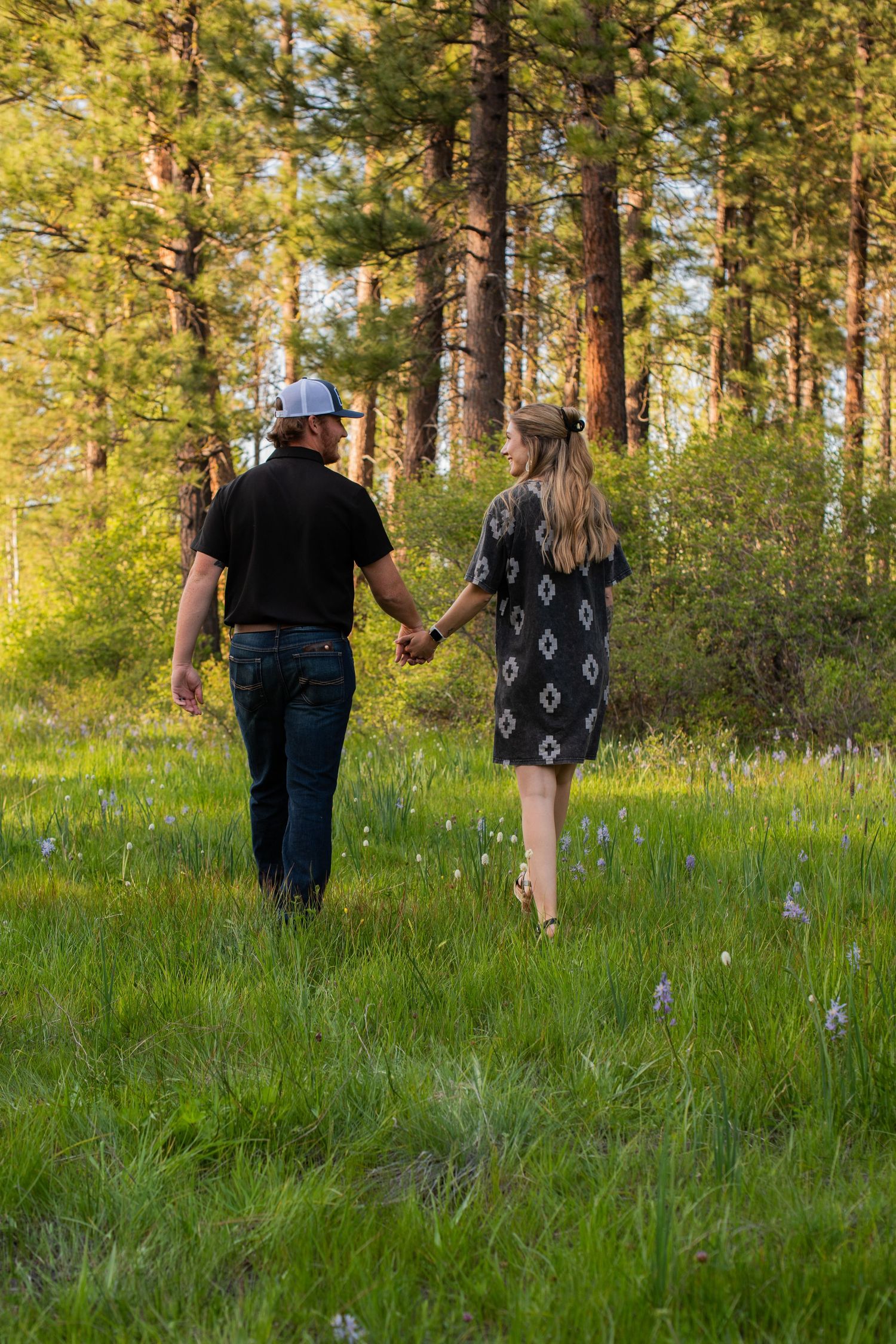 Couple walking hand in hand toward a pine forest through a green meadow with wildflowers at sunset.