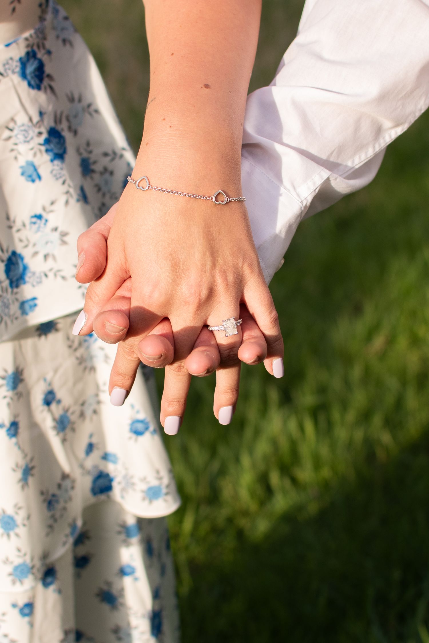 Close-up of engaged couple holding hands showing diamond engagement ring and delicate heart bracelet.