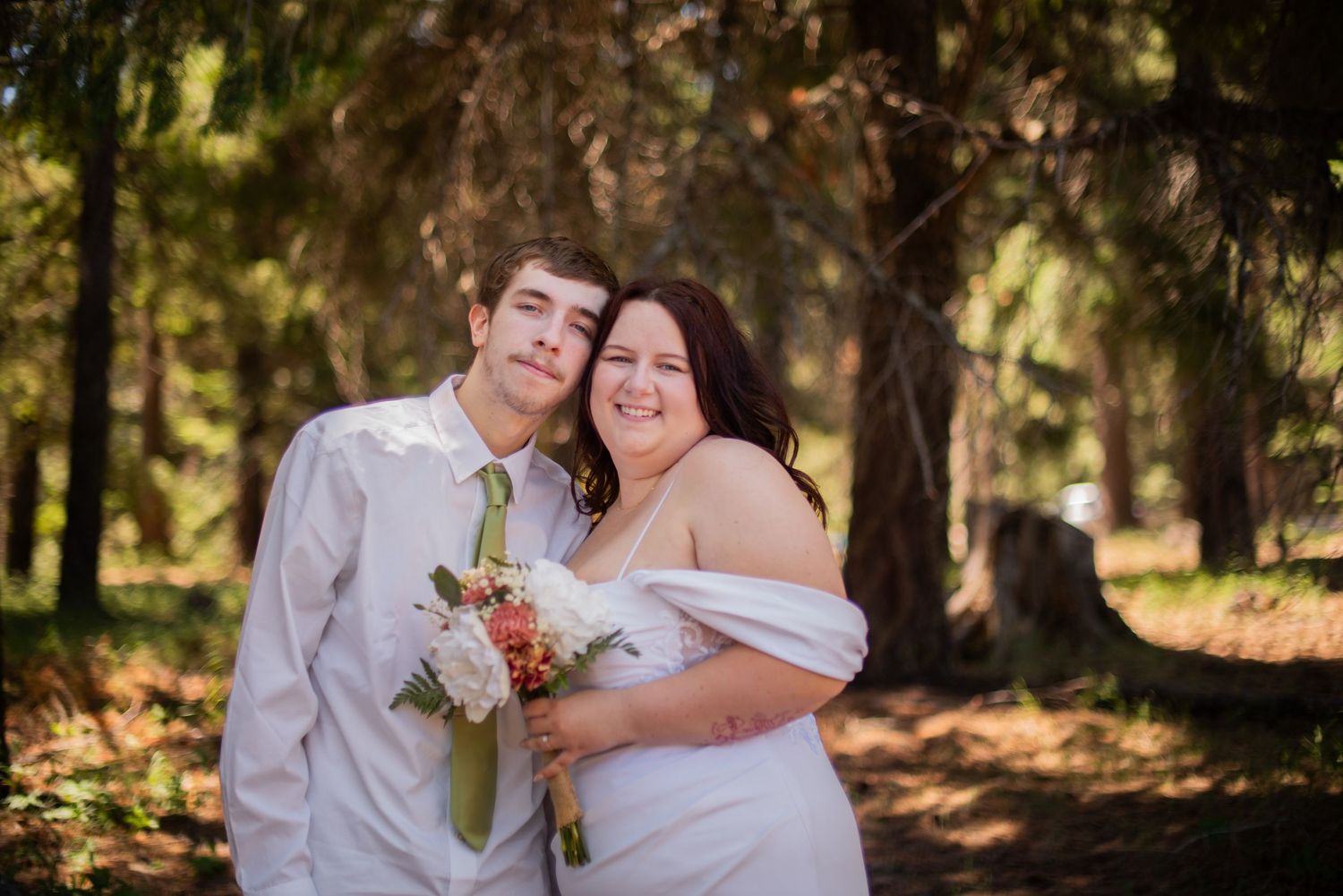 Smiling bride and groom embrace in a sunlit forest, bride holding a white and pink bouquet.