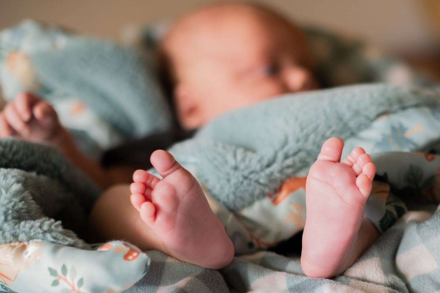 Tiny newborn baby feet peeking out from under a light blue blanket with a sleeping infant in background.