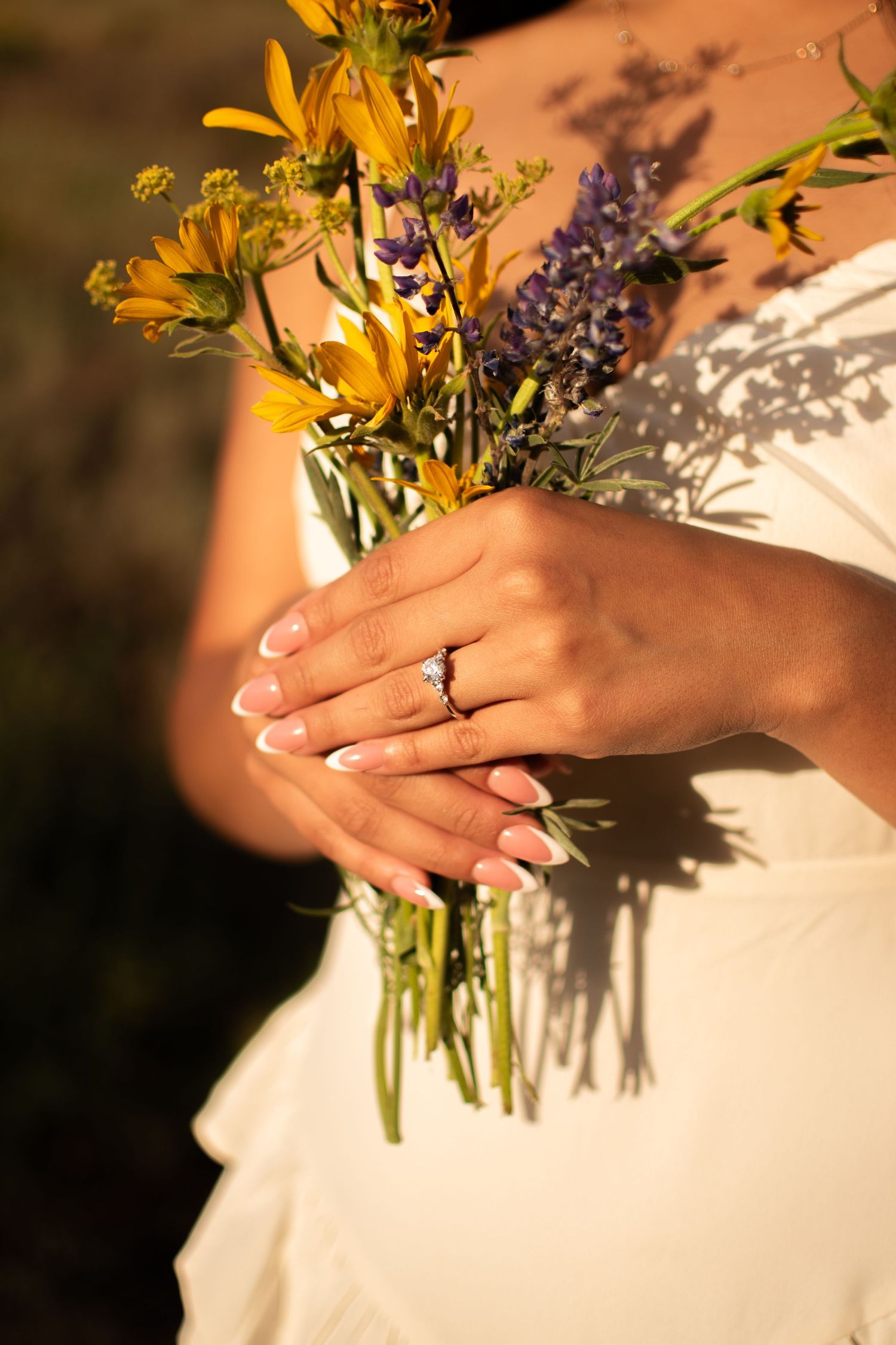 Bride holding wildflower bouquet with yellow and purple flowers, showcasing elegant French manicure and diamond engagement ring.