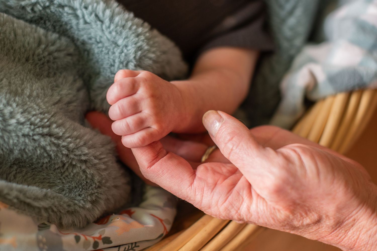 An elderly persons hand gently holding a newborn babys tiny fist on a soft teal blanket.