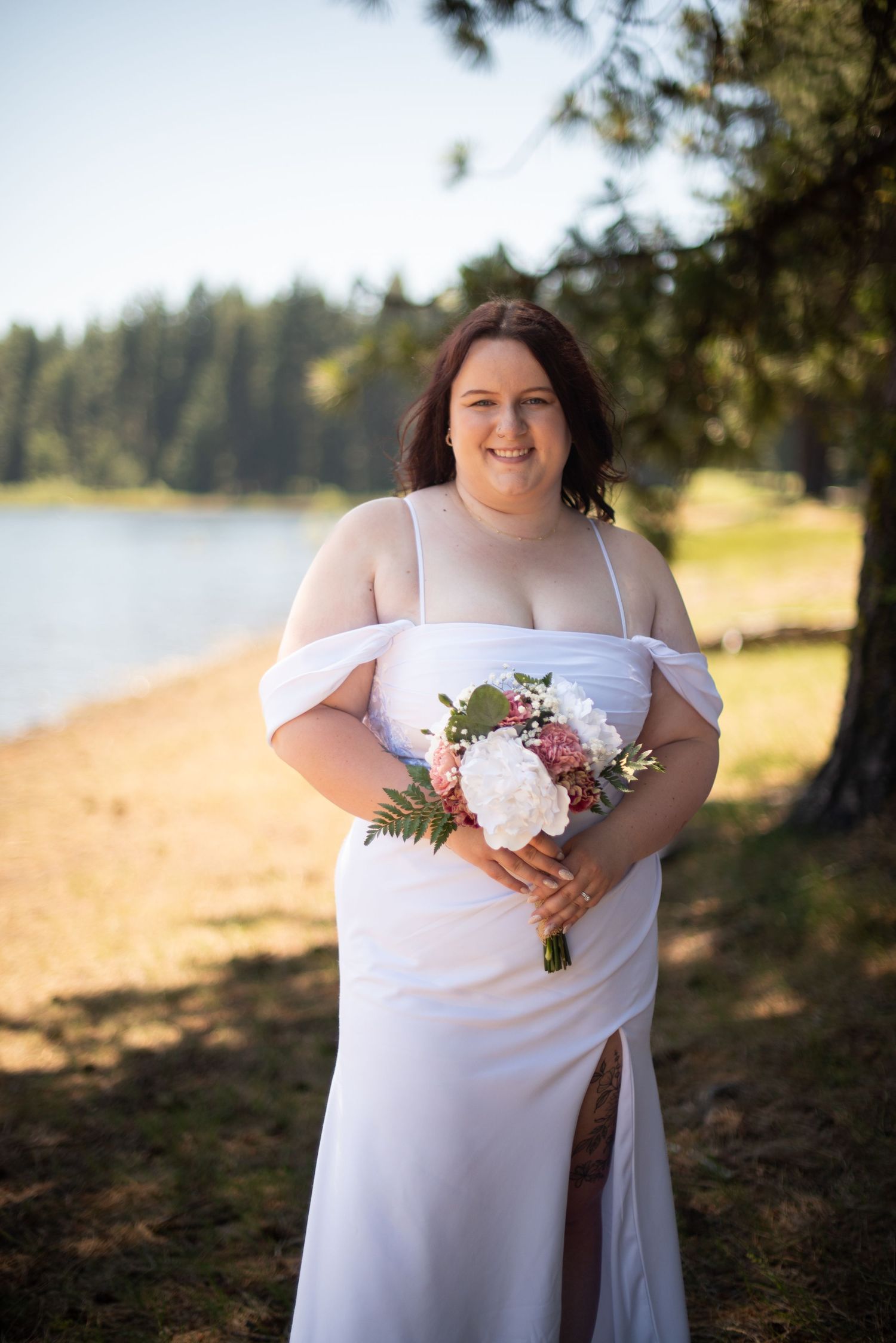 Bride in white off-shoulder wedding dress smiles holding bouquet by a lakeside pine tree at golden hour.
