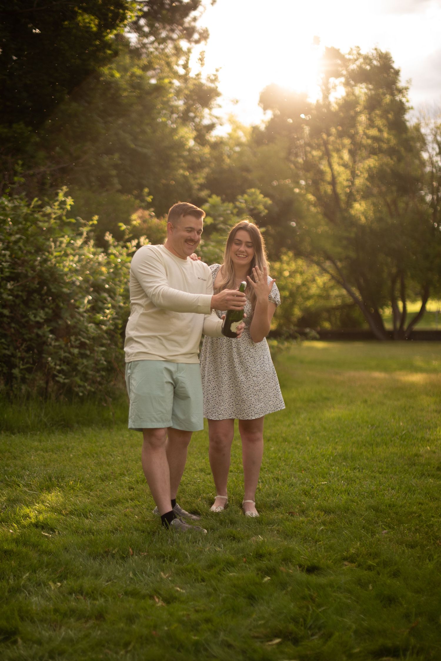 Couple celebrating an engagement in a park, woman showing off ring while man pours champagne in golden sunlight.