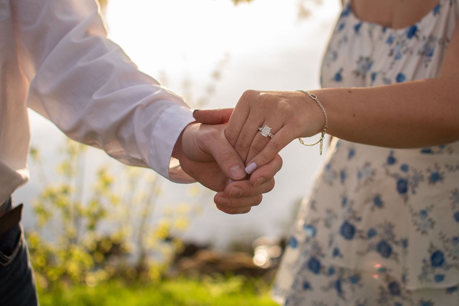 Couple holding hands outdoors, woman wearing a diamond engagement ring, romantic proposal moment in natural light.