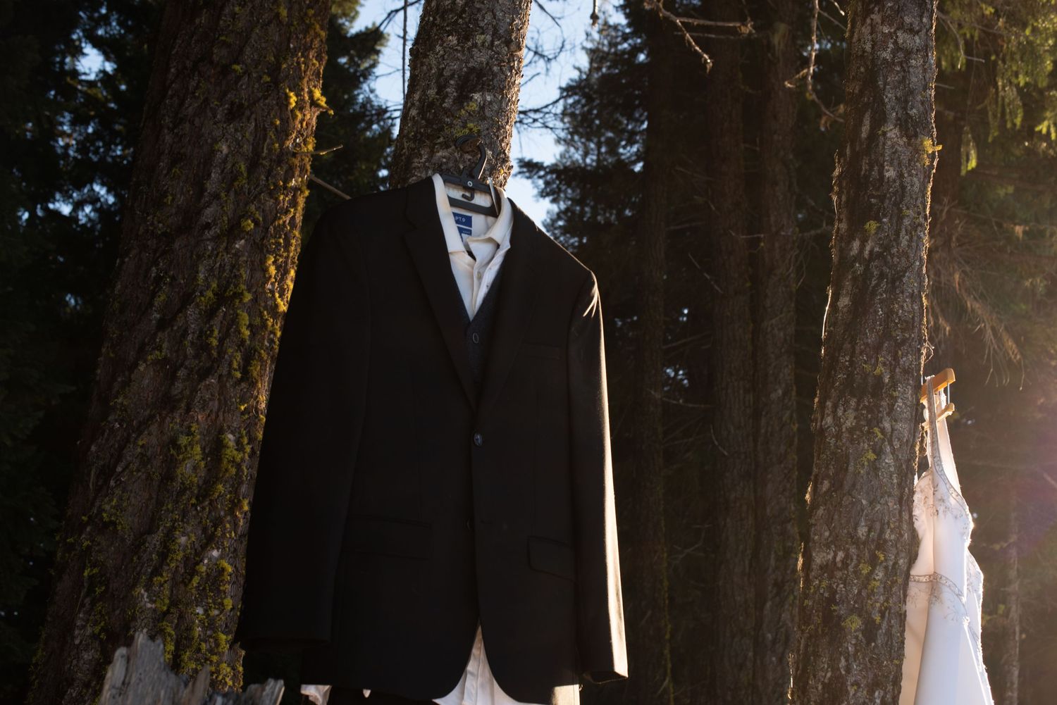 Black suit jacket hanging on a hanger between trees in a forest, with a white dress visible in the background.