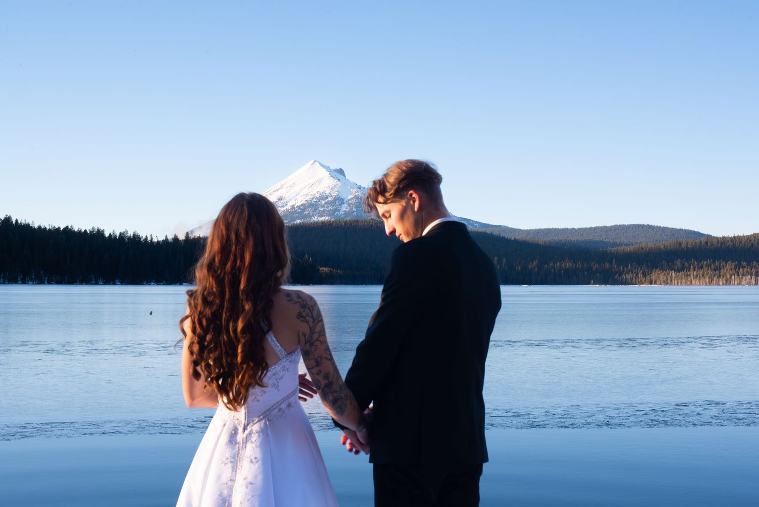 Bride and groom hold hands facing a stunning snow-capped mountain reflected in a calm lake.