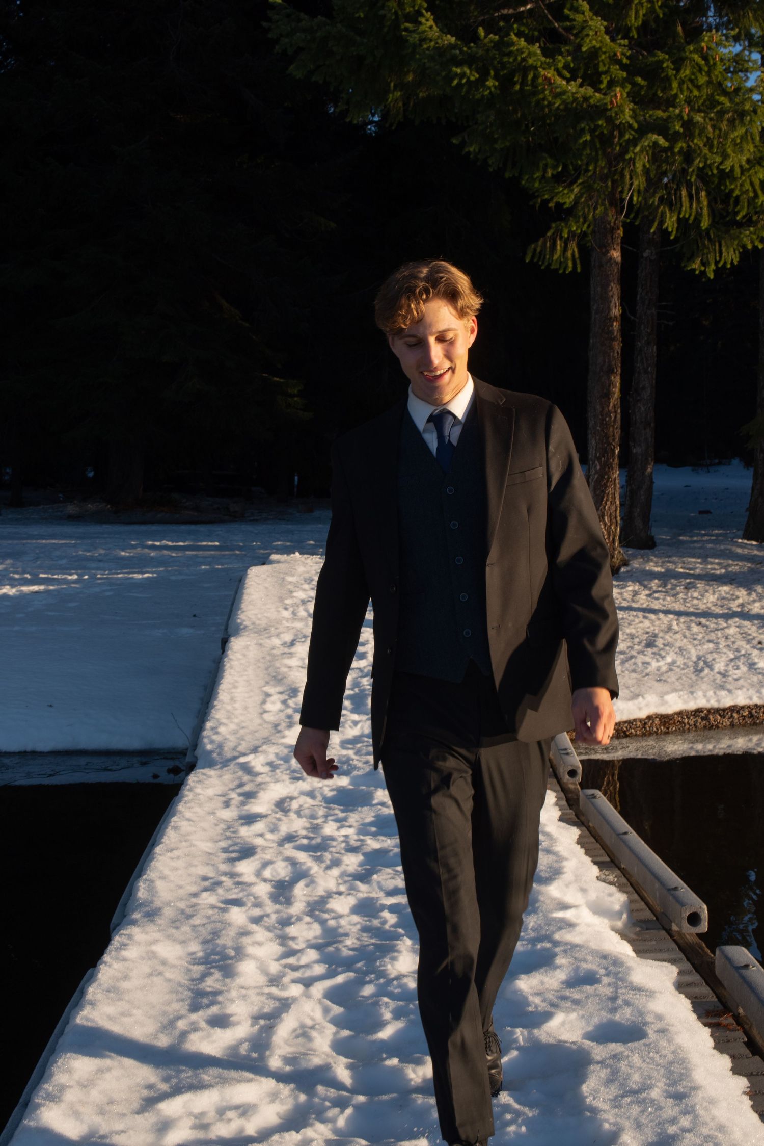 Groom in dark suit walking on snow-covered dock surrounded by pine trees in golden light.