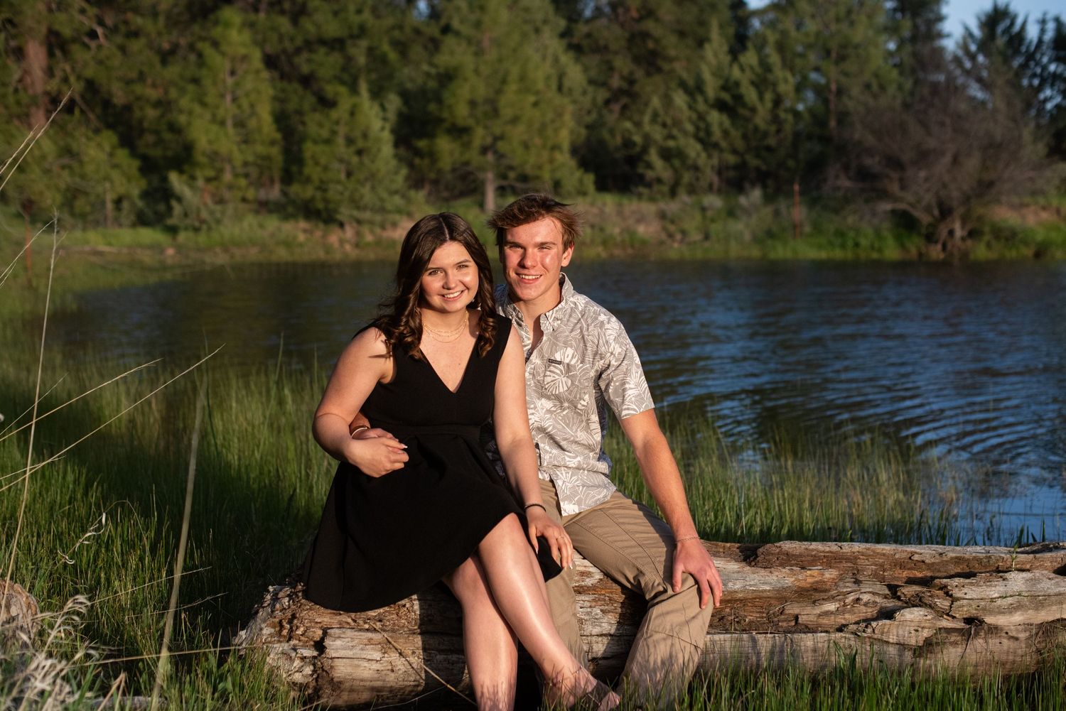 A smiling couple sits together on a log near a peaceful river surrounded by lush green trees and grass.