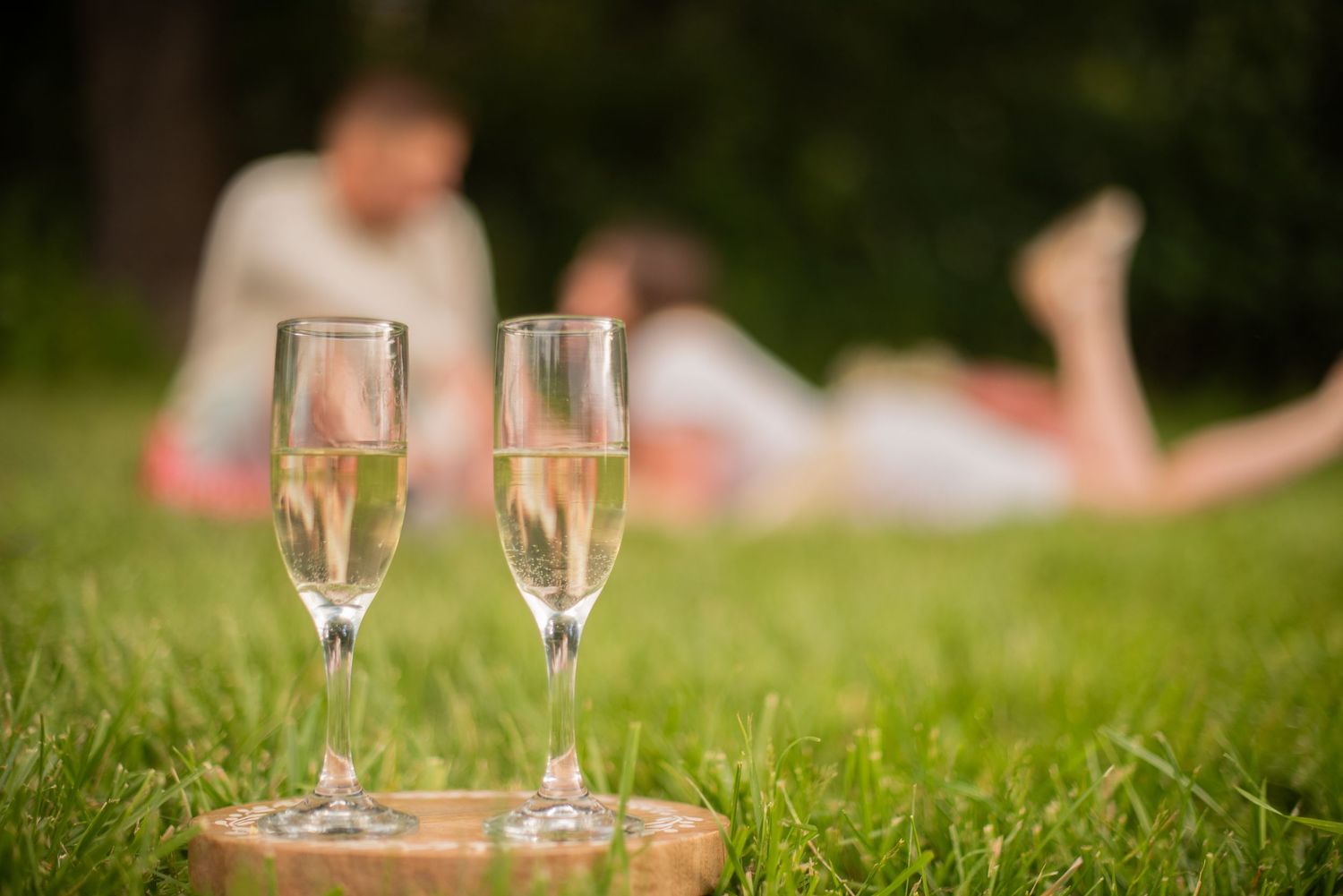 Two champagne flutes on grass with couple relaxing in soft focus background at outdoor picnic.