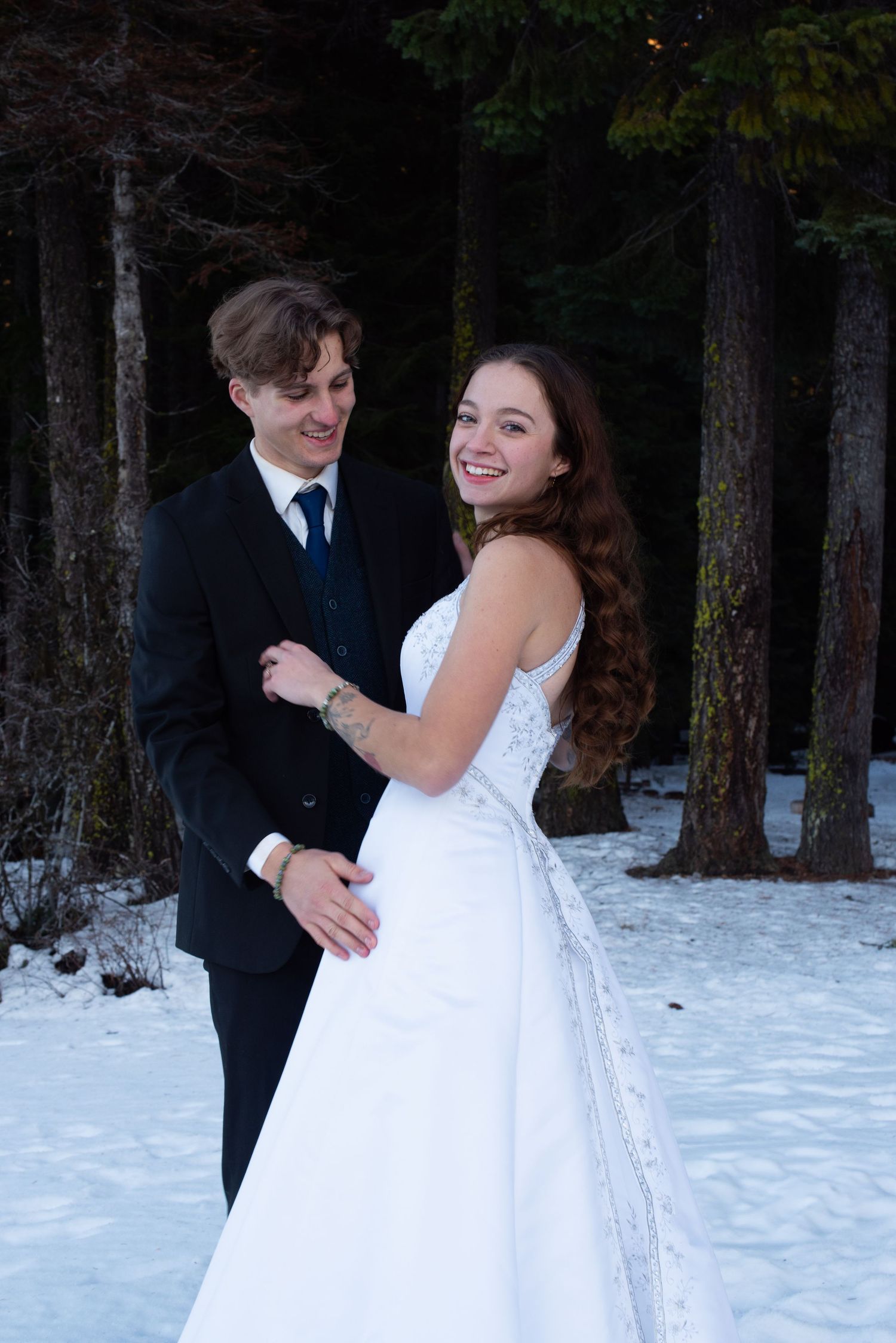 Smiling bride in white gown and groom in black suit embracing in snowy forest setting.