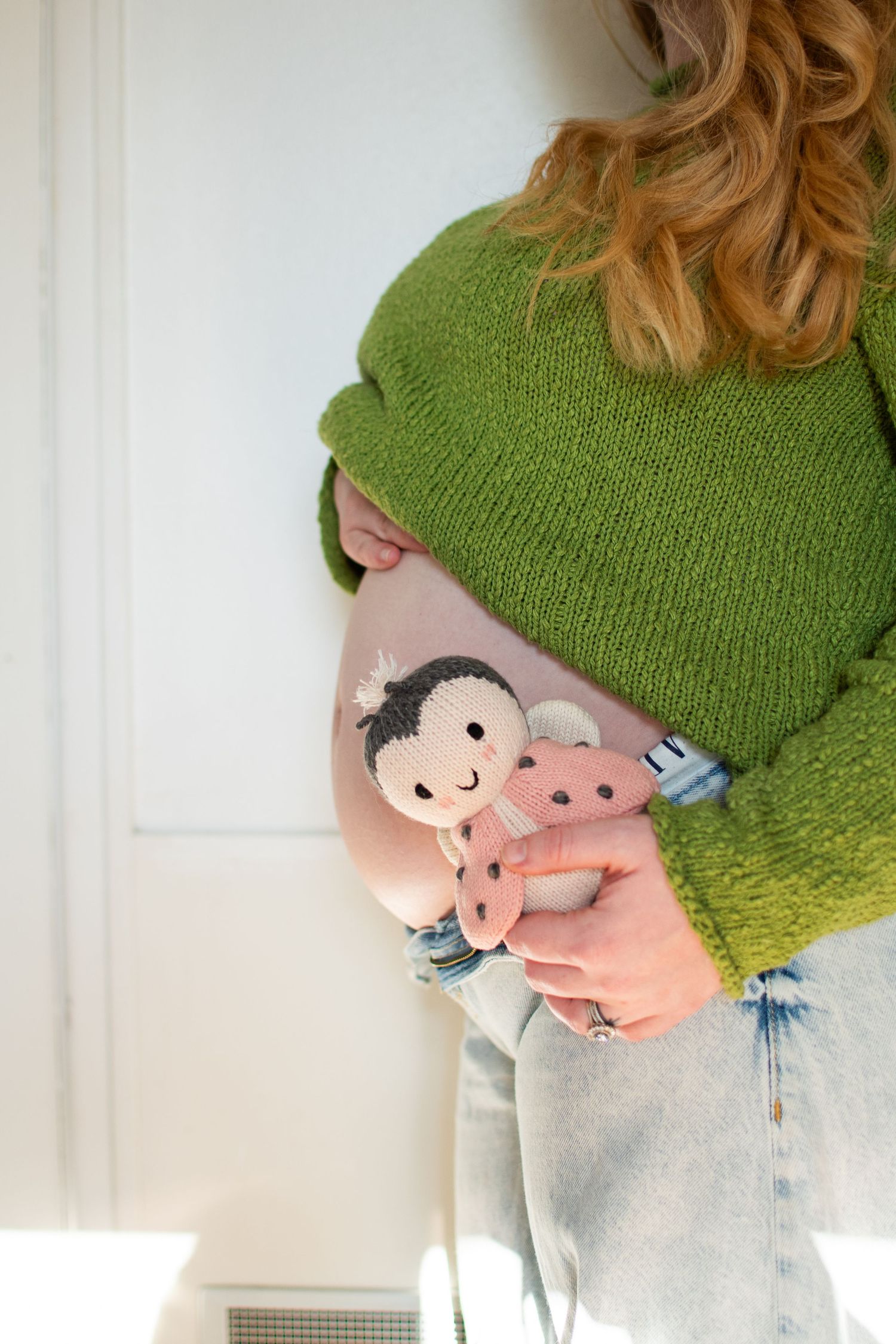 Pregnant woman in green knit sweater holding a pink ladybug stuffed doll against her baby bump.