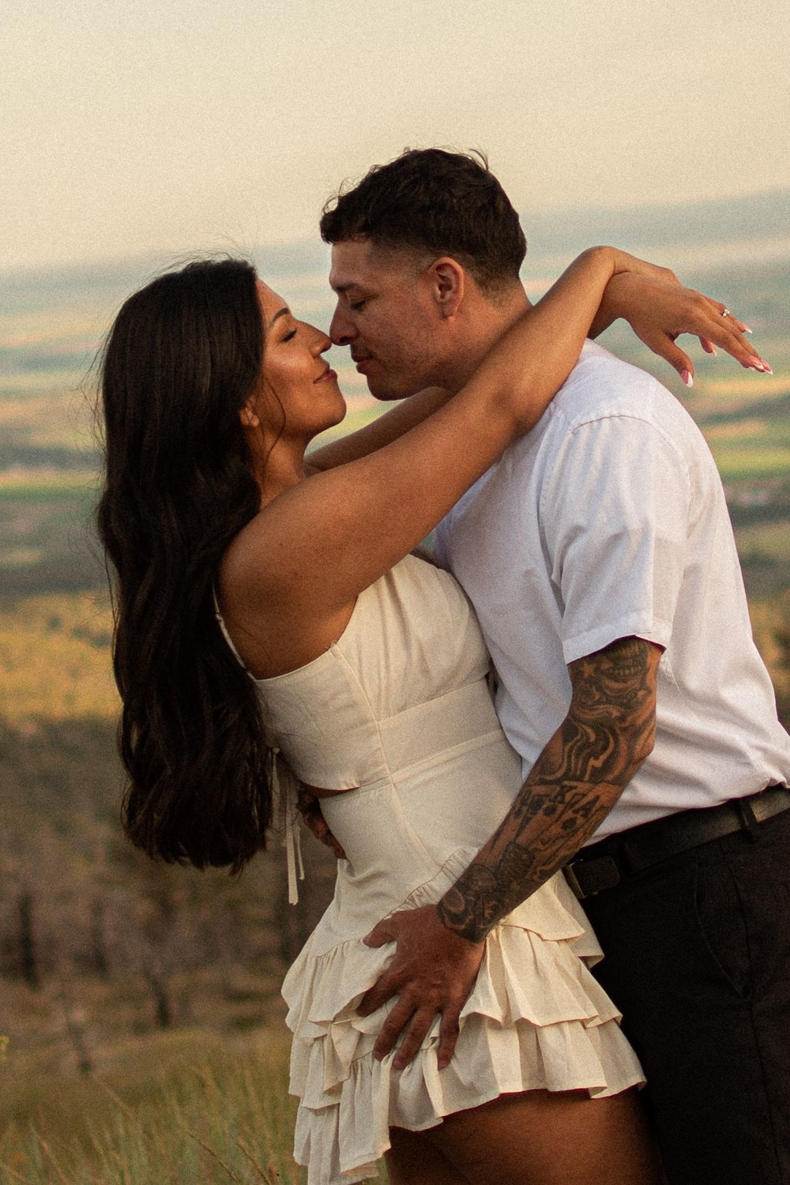 Couple in white outfits embracing on a hilltop, woman in ruffled mini dress, man with tattoos, scenic valley backdrop.