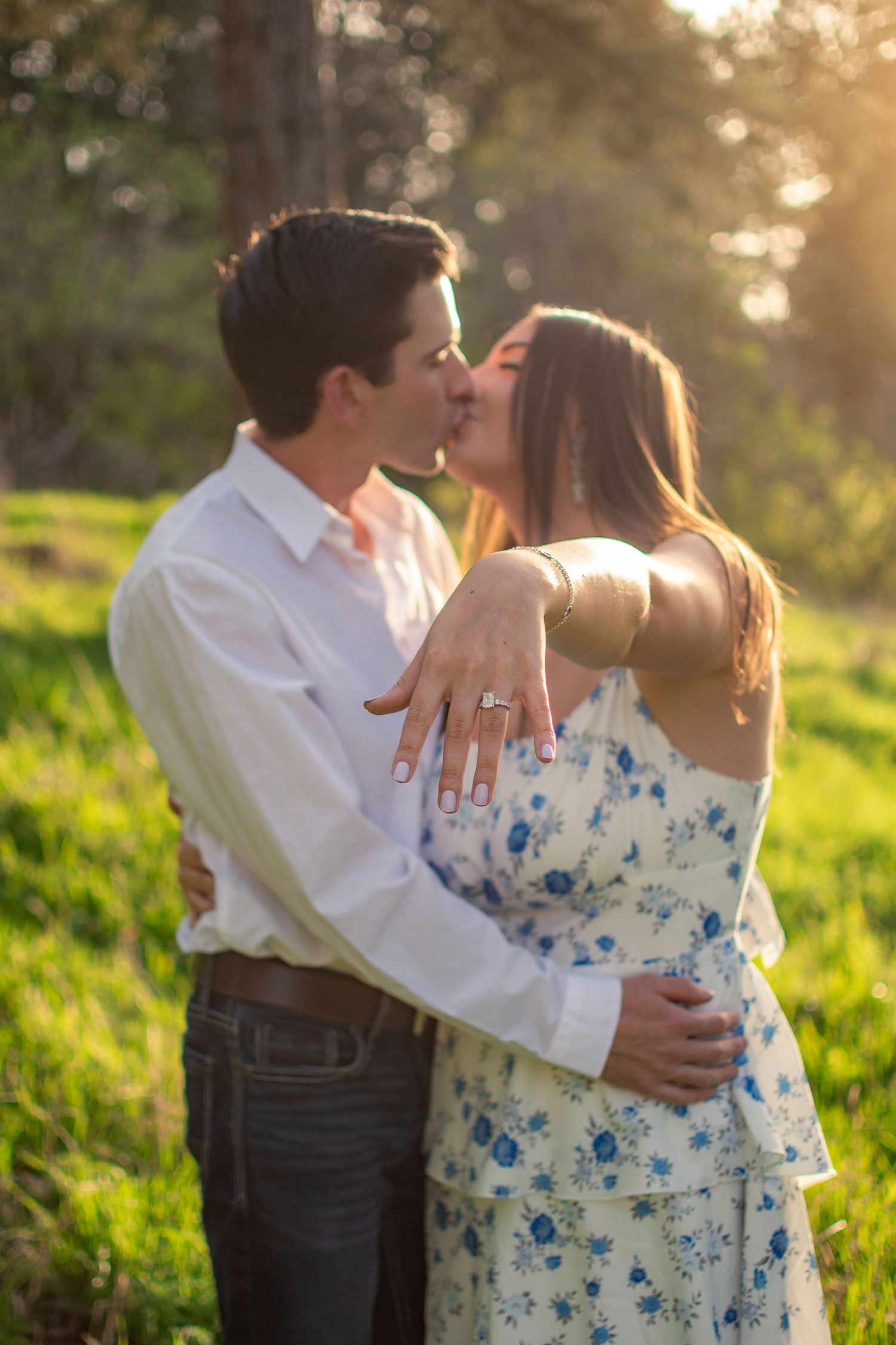 Couple kissing outdoors at golden hour, woman displaying engagement ring while embracing in a sunlit green field.