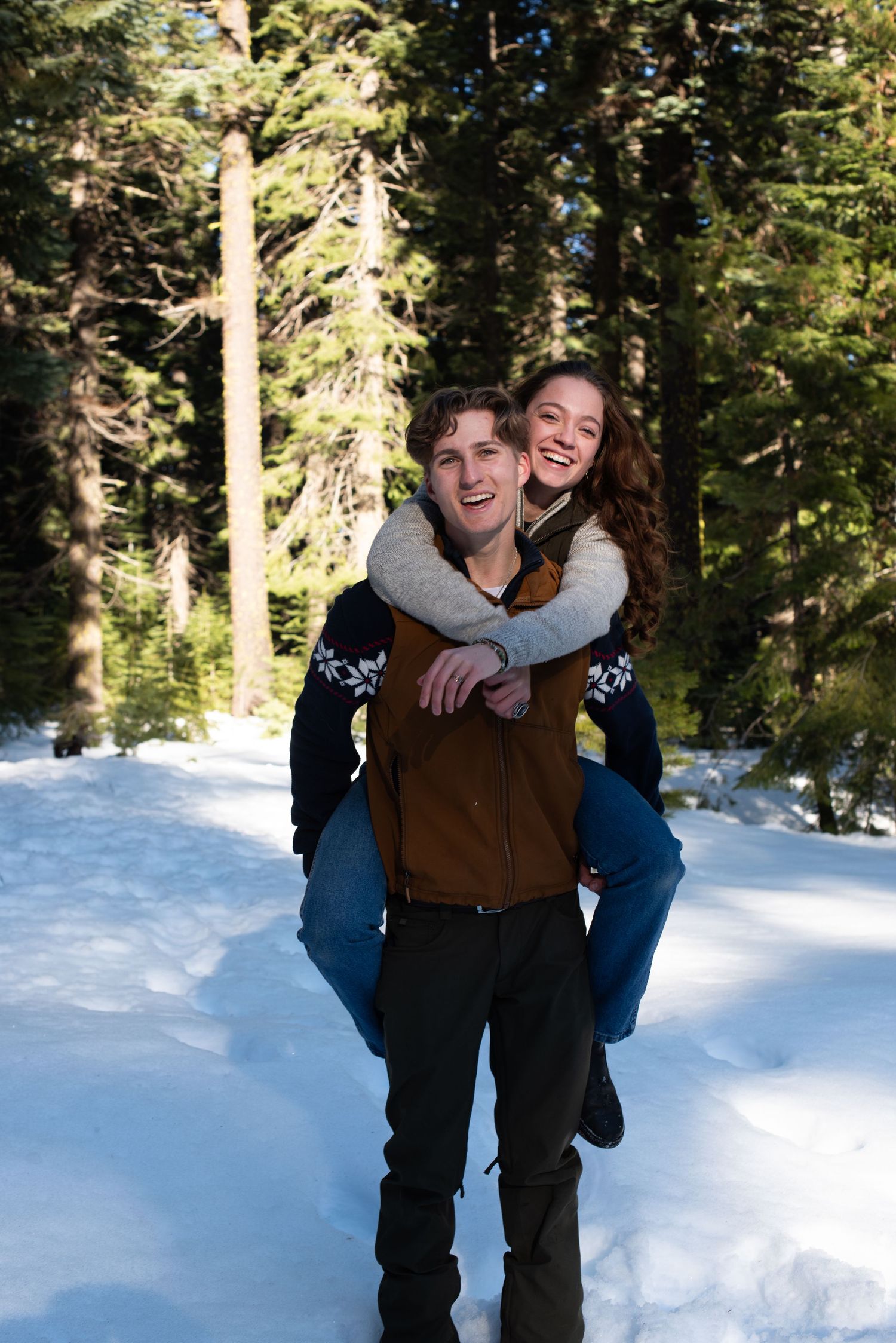 Couple laughing in snowy forest, man giving woman piggyback ride among tall pine trees in winter.