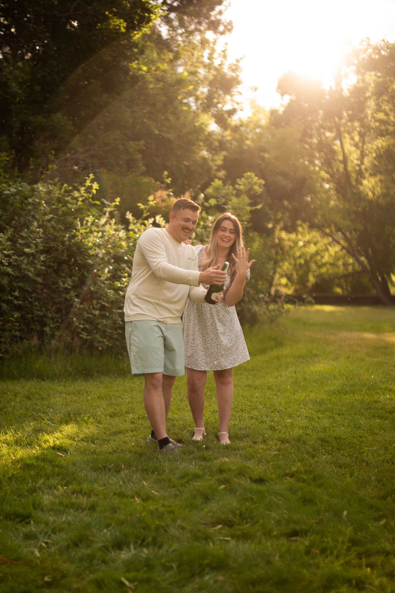 Happy couple sharing a bottle of champagne in a sunlit green park during golden hour.