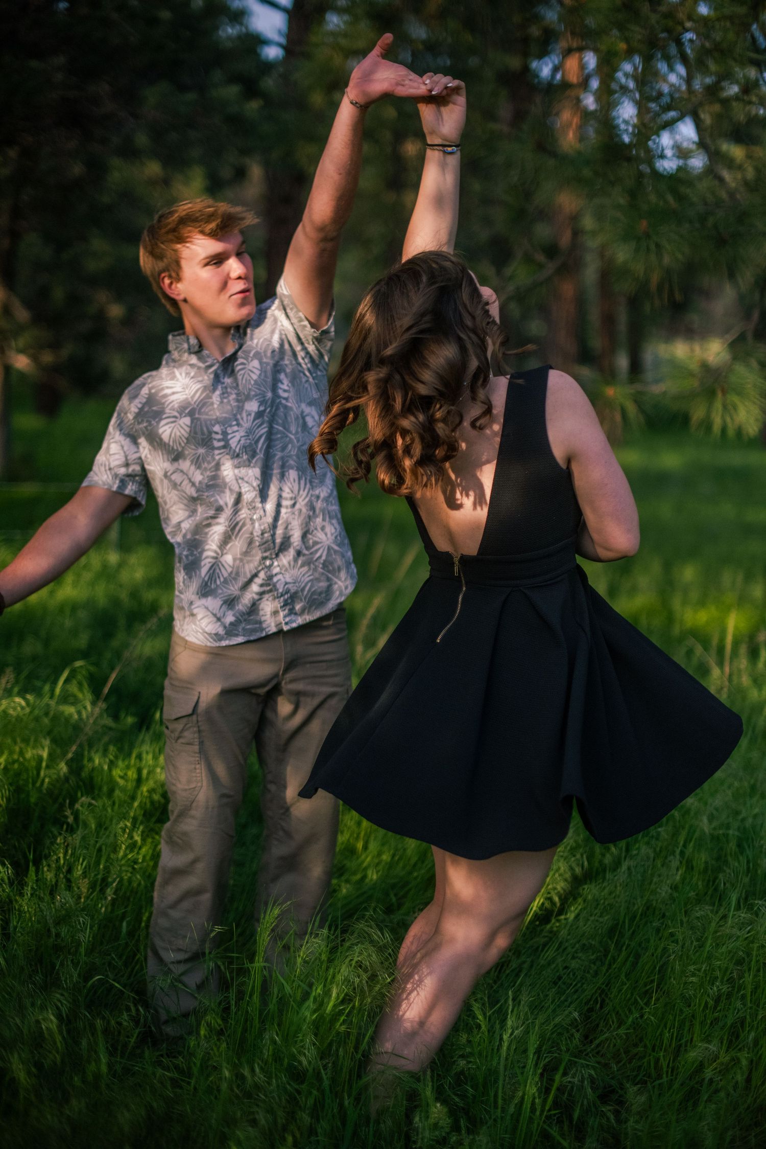 A couple dances together in a lush green meadow surrounded by tall pine trees in golden sunlight.